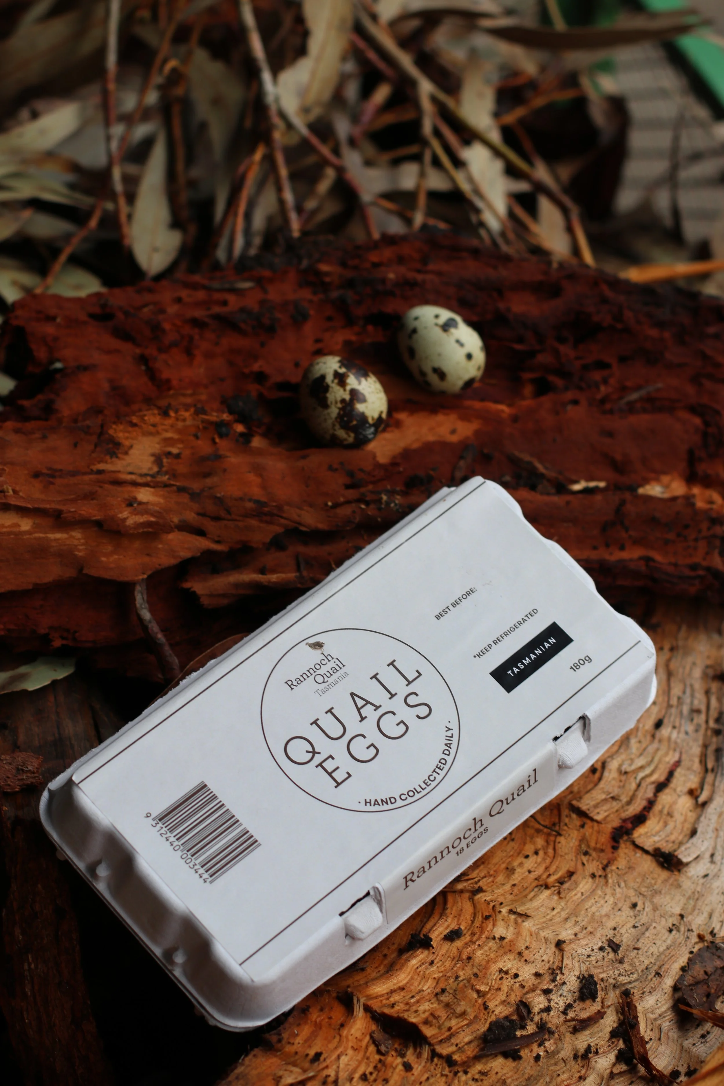 A white carton of Quail Eggs from Rannoch Quail in Tasmania, placed on a rough wooden surface with scattered leaves and twigs in the background. Two speckled quail eggs are resting on the wood next to the carton.