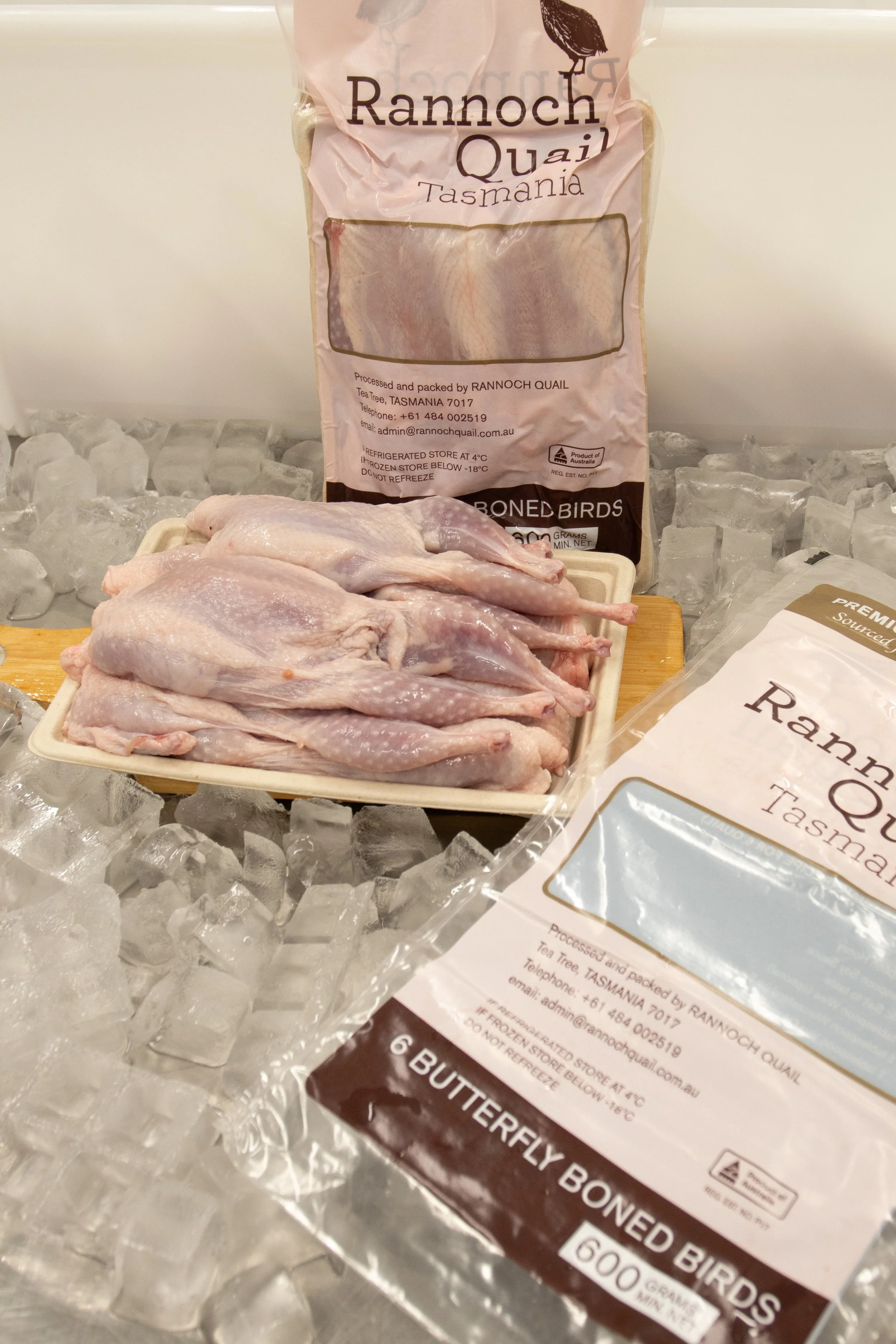 Frozen packages of Rannoch Quail Tasmania and bone-in bird parts on ice, with a tray of raw quail in front.