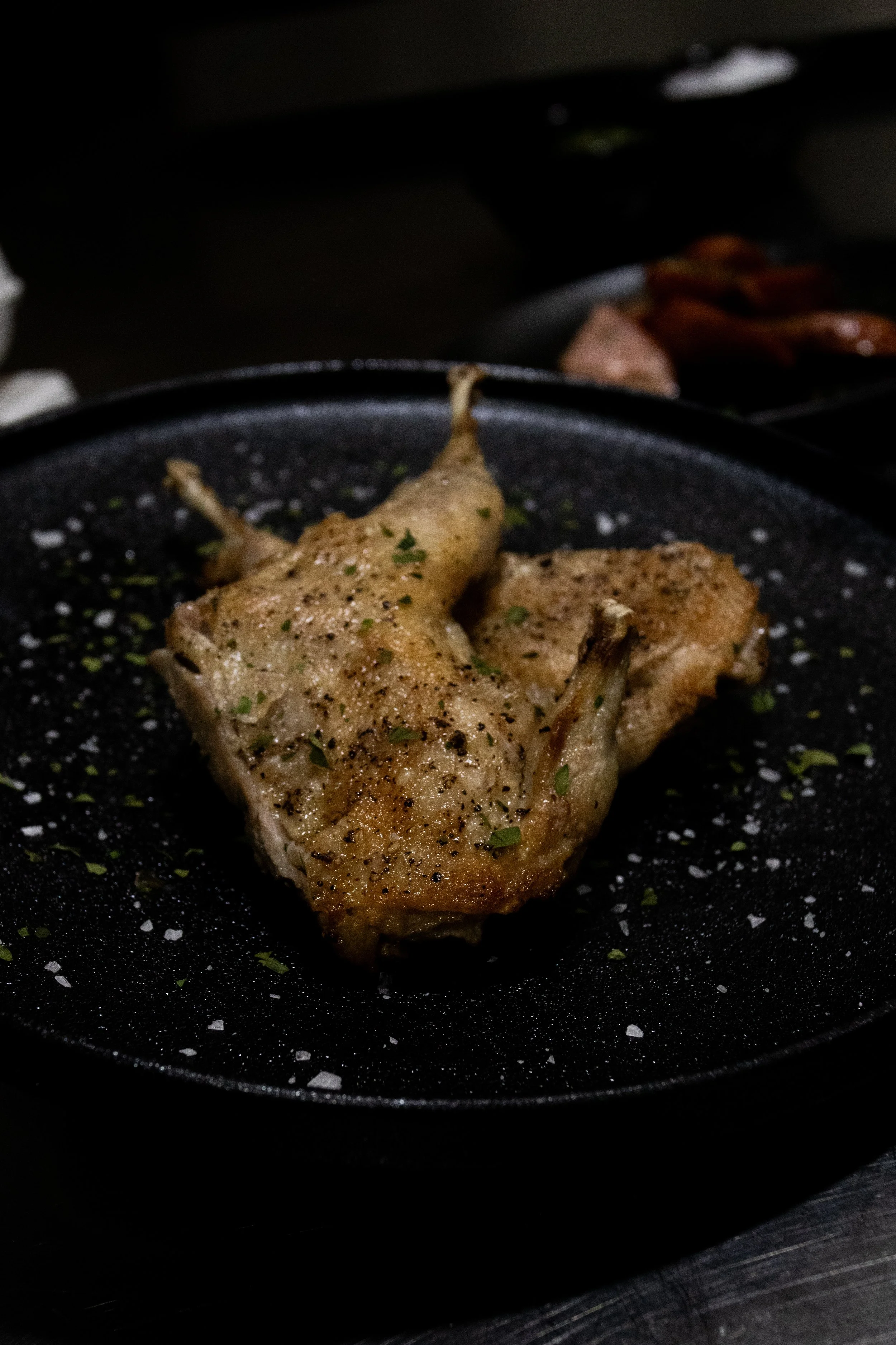 Cooked chicken thigh seasoned with herbs and black pepper, on a black skillet with salt flakes, with other ingredients faintly visible in the background.