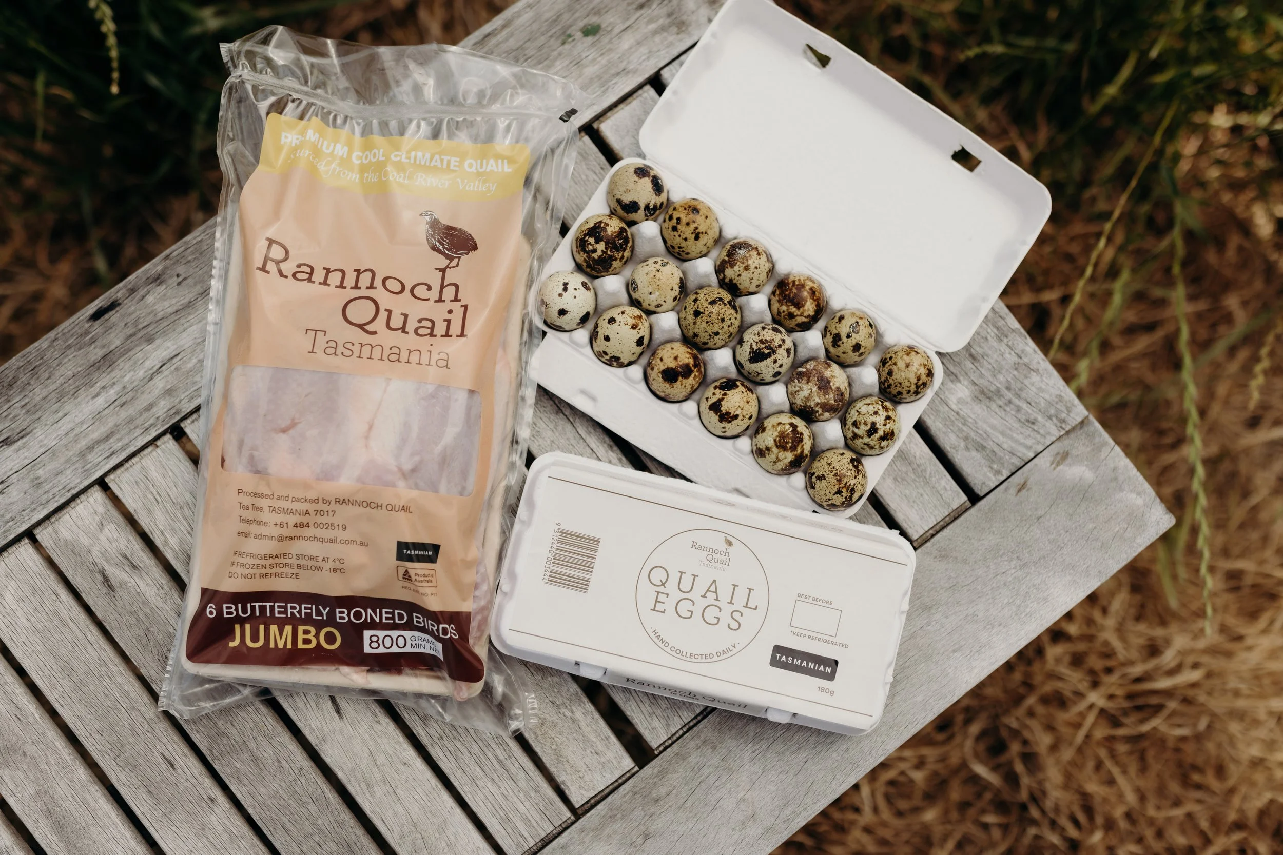 Packet of Rannoch Quail Tasmania jumbo butterfly boned quail birds and a carton of quail eggs on a weathered wooden table outdoors.