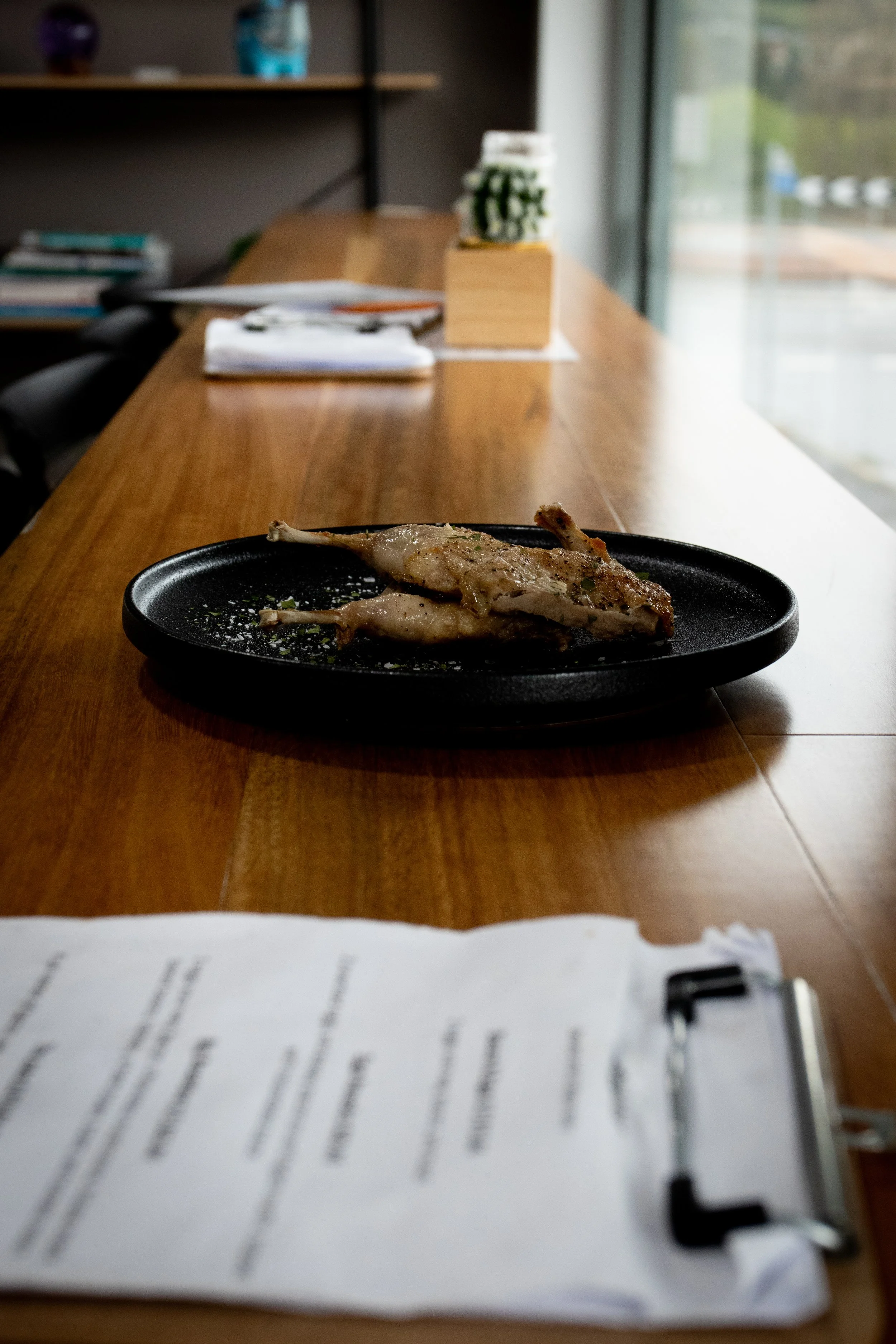 A plate with a cooked chicken leg quarter garnished with herbs on a wooden table in a bright room with large windows.