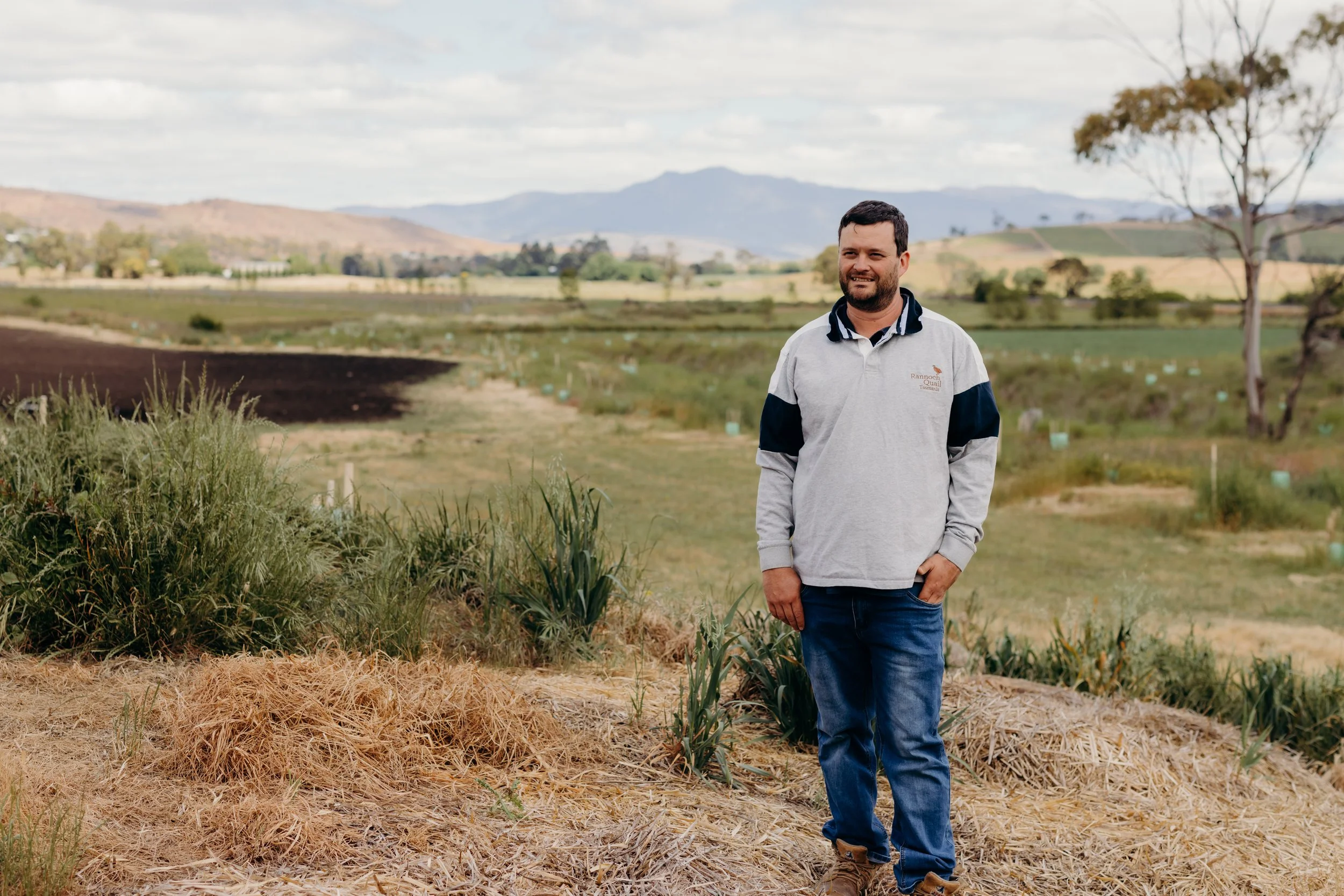 A man standing outdoors on a farm or agricultural field with mountains in the background, wearing a light gray and navy hoodie and jeans.