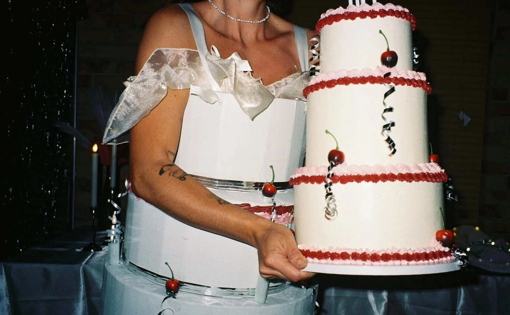 A woman holding a three-tiered white birthday cake decorated with cherries and black and silver curl ribbons. She's wearing a silver dress with ruffle sleeves and a pearl necklace. The background features a dark wall with some lights and decorations.