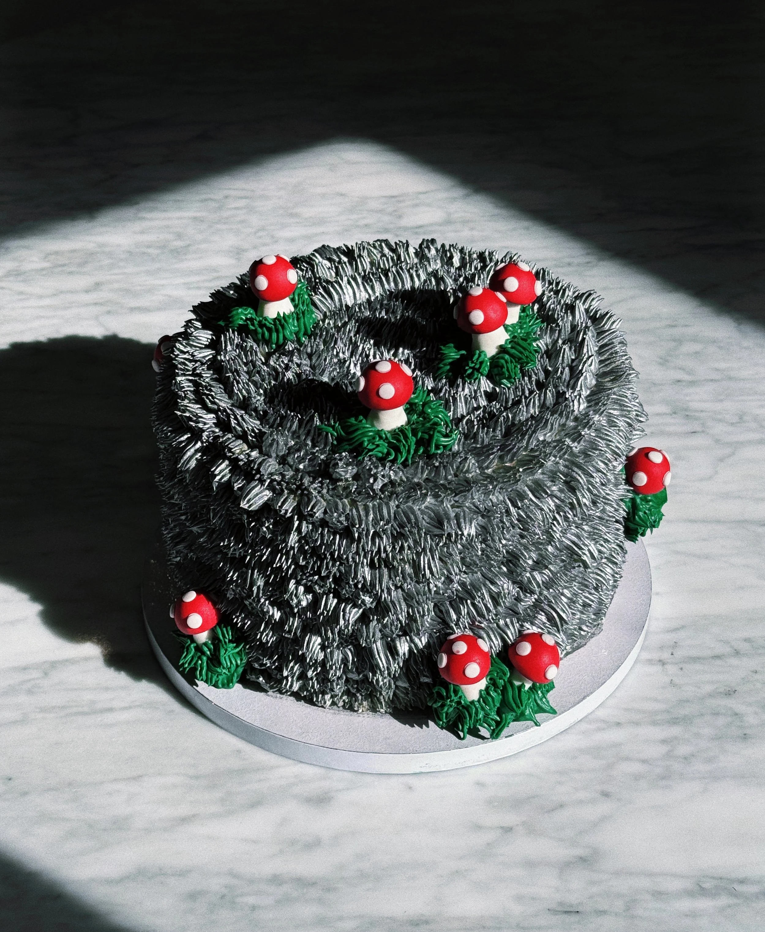 Decorative cake with silver icing, green icing leaves, and red mushrooms with white spots, on a white round tray.
