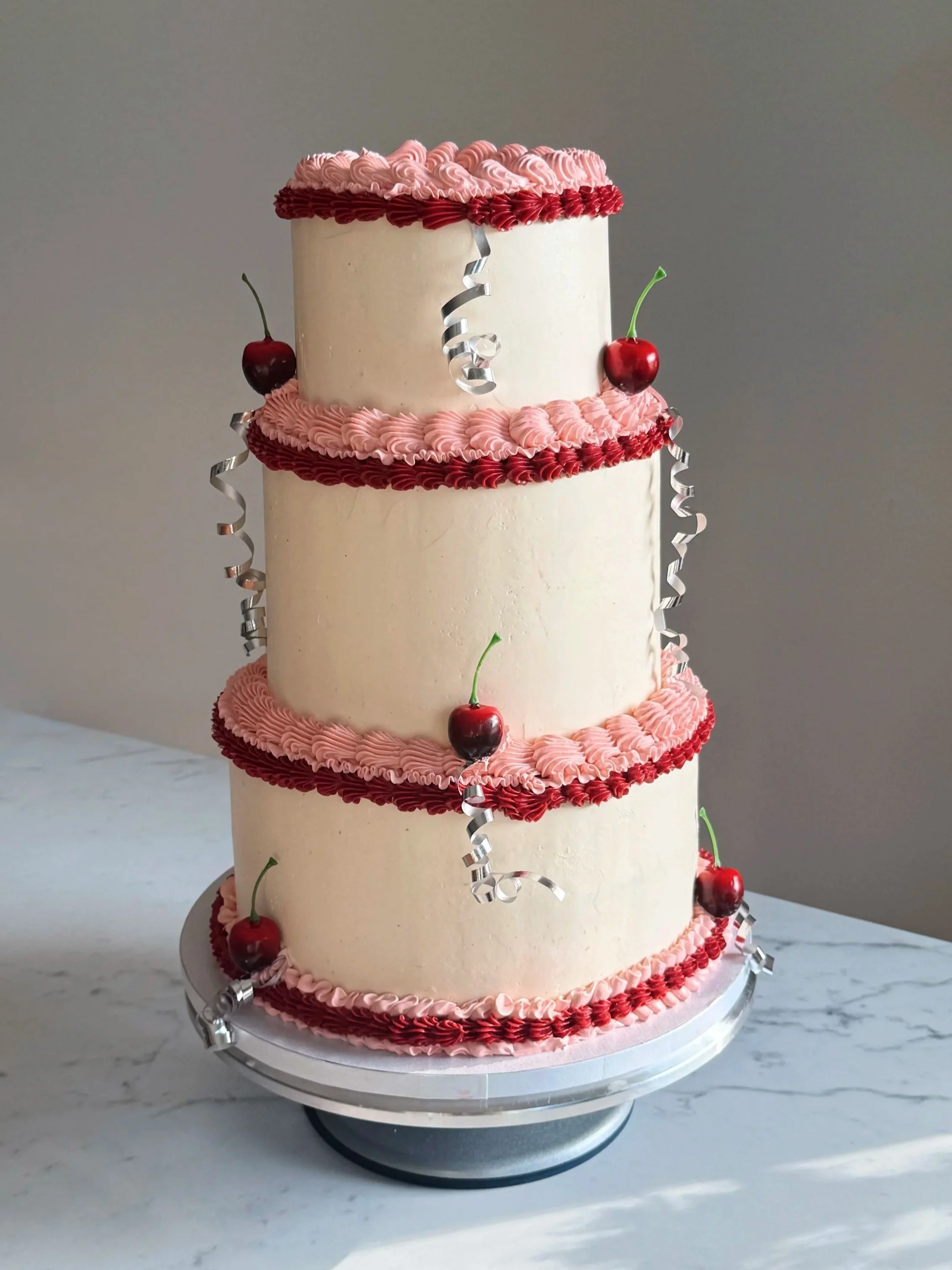 A three-tiered white cake decorated with pink and red frosting, topped with cherries and silver curly ribbons, on a marble surface.