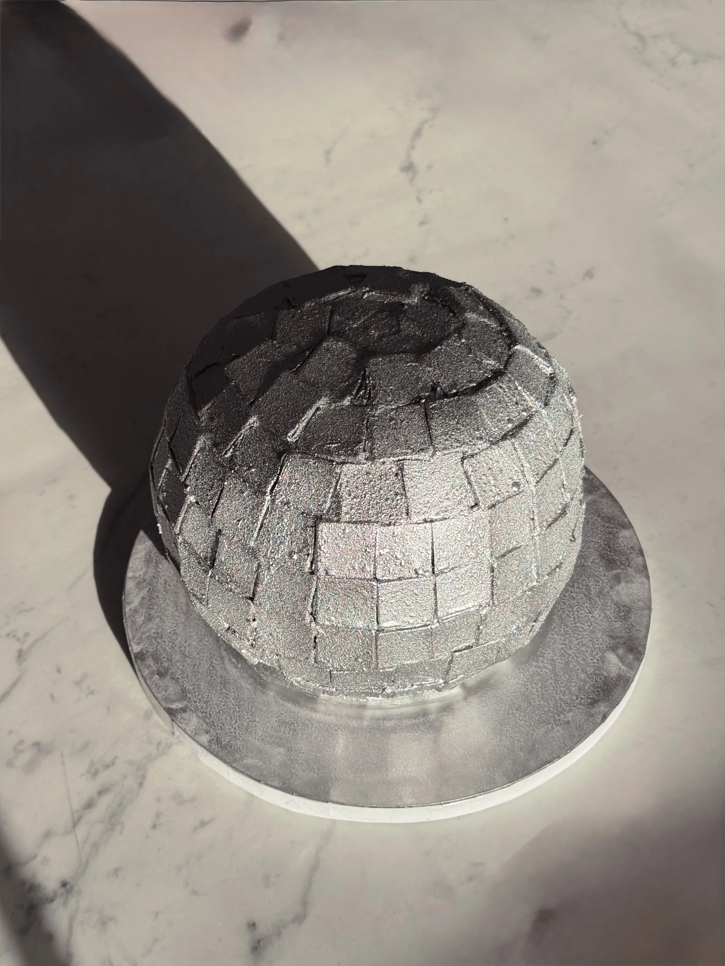 A cylindrical object with a textured, crackled silver surface placed on a reflective surface. The object is casting a shadow on the surface.