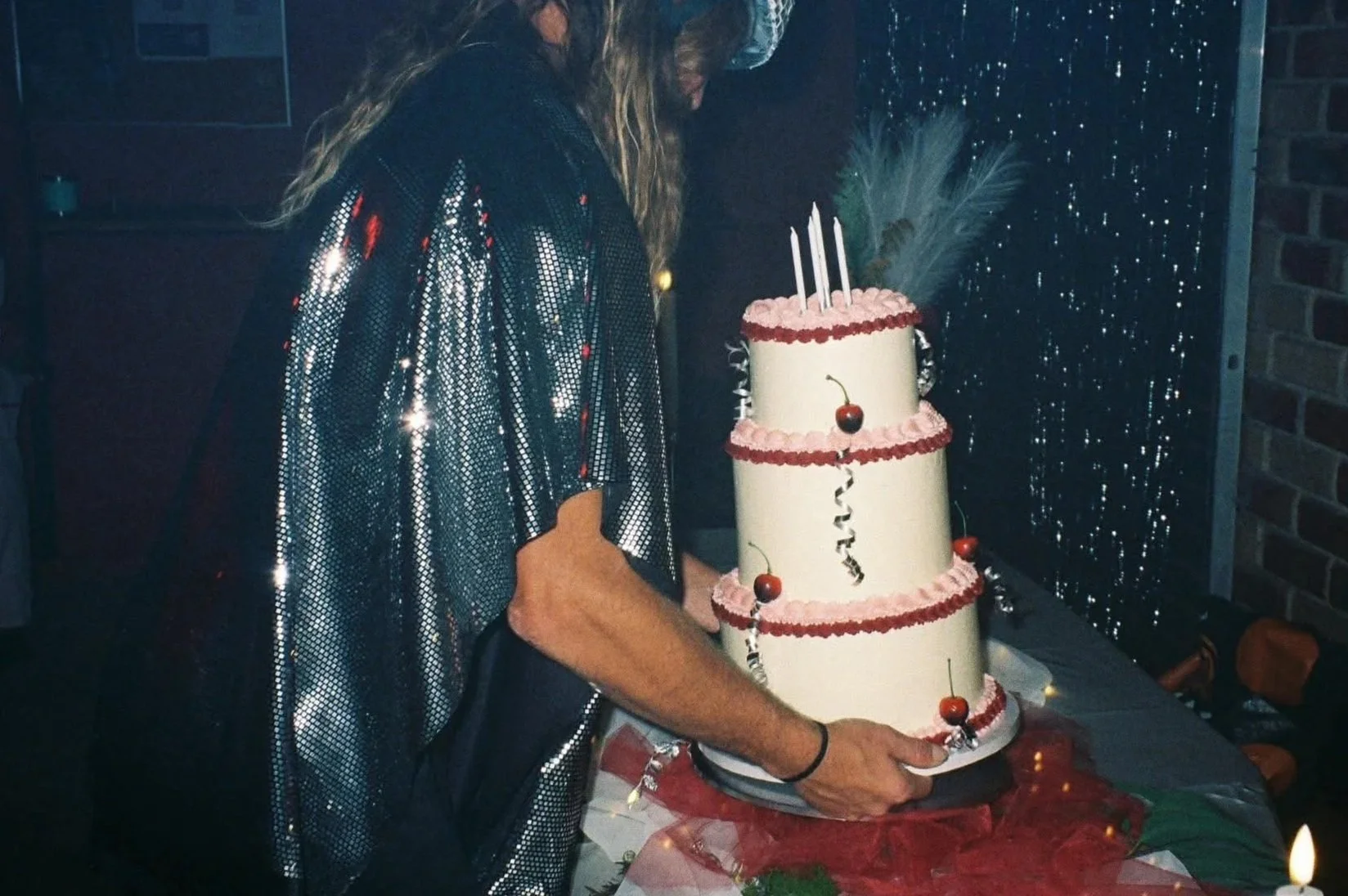 Person in a shiny, patterned shirt carrying a three-tiered birthday cake decorated with cherries, pink frosting swiss buttercream, and candles in a festive birthday setting.