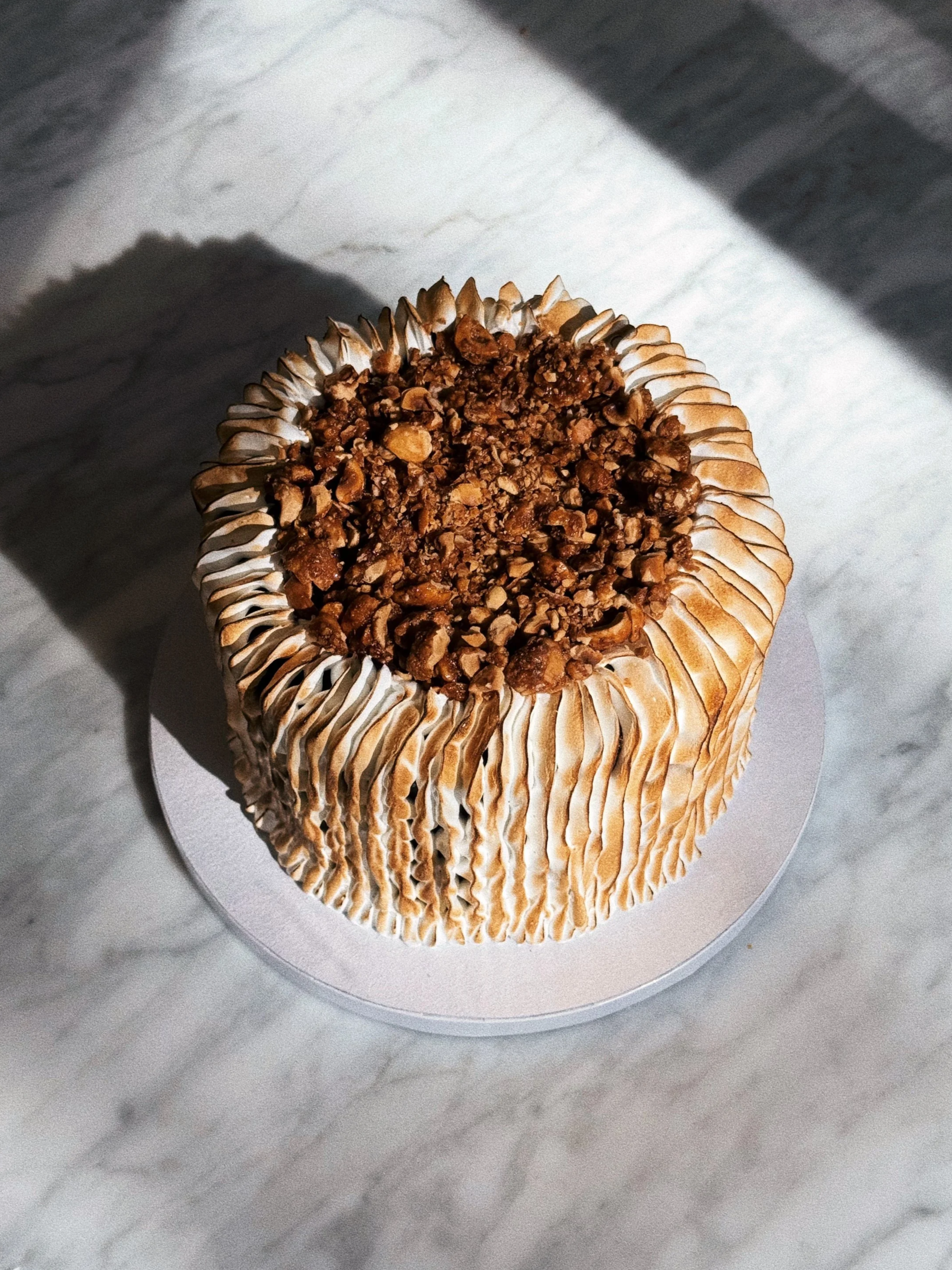A cake with burnt Italian meringue and chopped caramelised hazelnuts on top, placed on a white plate on a marble surface.