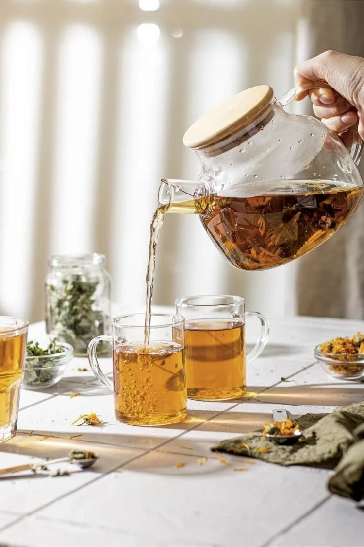 A person is pouring tea from a glass teapot with a wooden lid into glass mugs on a white tiled table. There are additional teacups, a jar with herbs, and some scattered herbs and flowers on the table, with a light background.