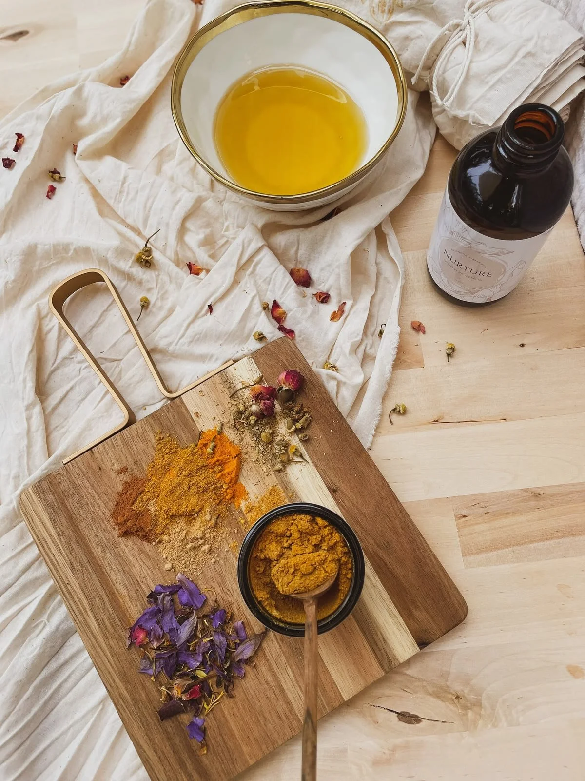 Various dried herbs and spices on a wooden cutting board, a bowl of yellow turmeric powder, a dark bottle labeled 'Nurture', a glass bowl with yellow spice, a spoonful of turmeric powder, a white cloth with scattered dried rosebuds, and purple flower petals, all arranged on a light wooden surface.