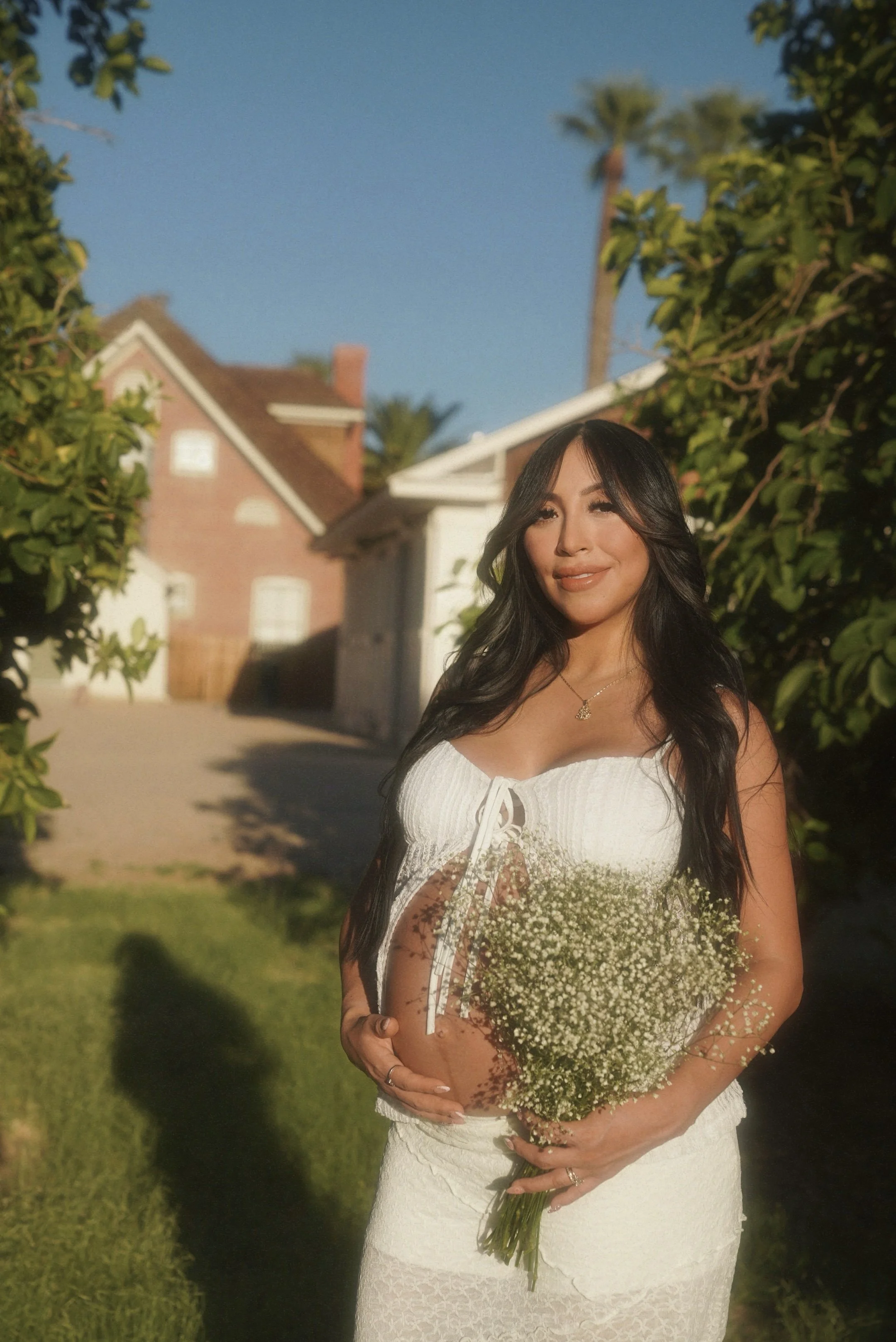 A pregnant woman in a white dress holding a bouquet of small white flowers, standing outdoors in front of a house with a garden and some trees.