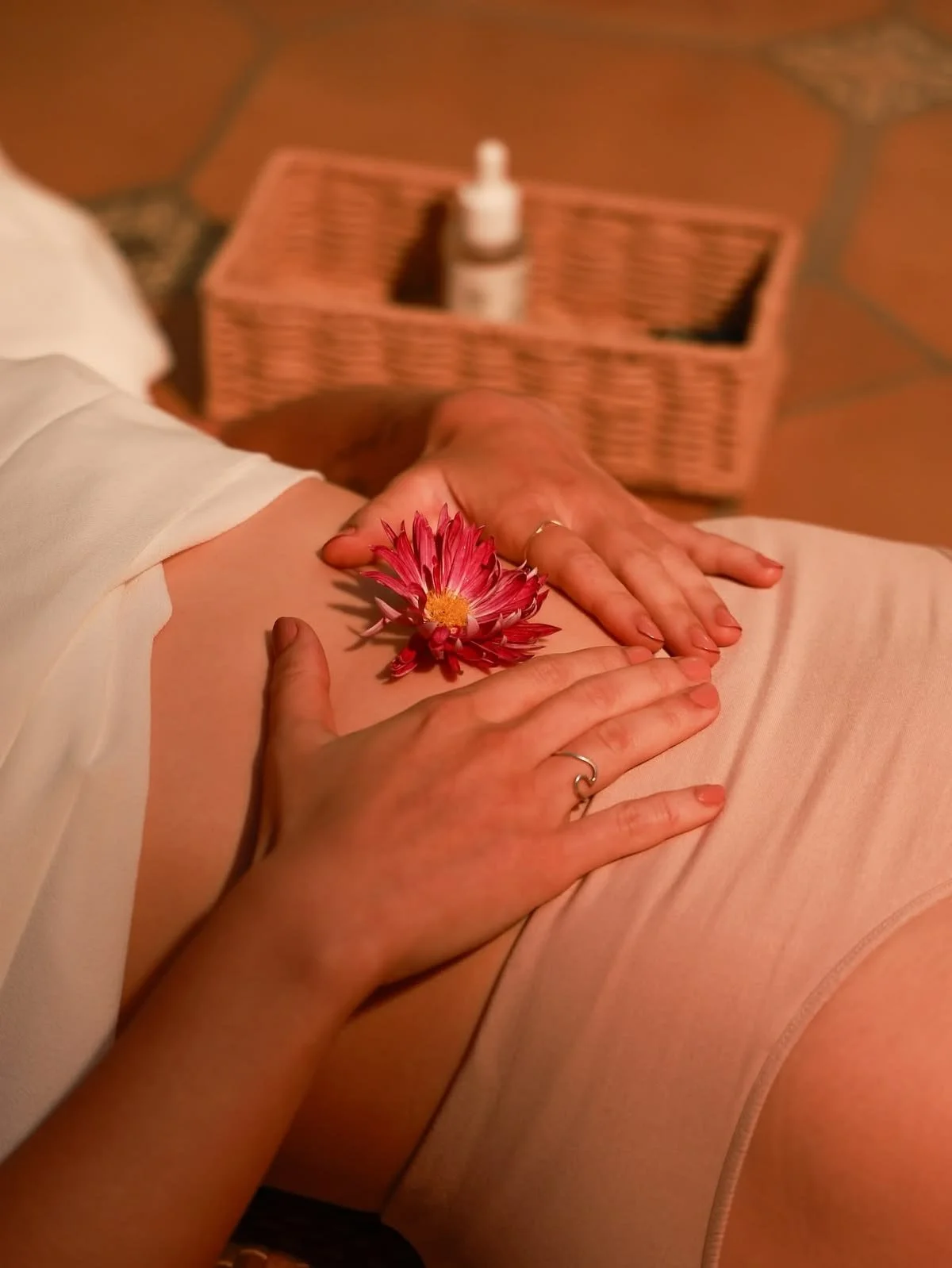 A person receiving a massage, with a pink flower on their bare skin. There is a basket in the background with a bottle inside.