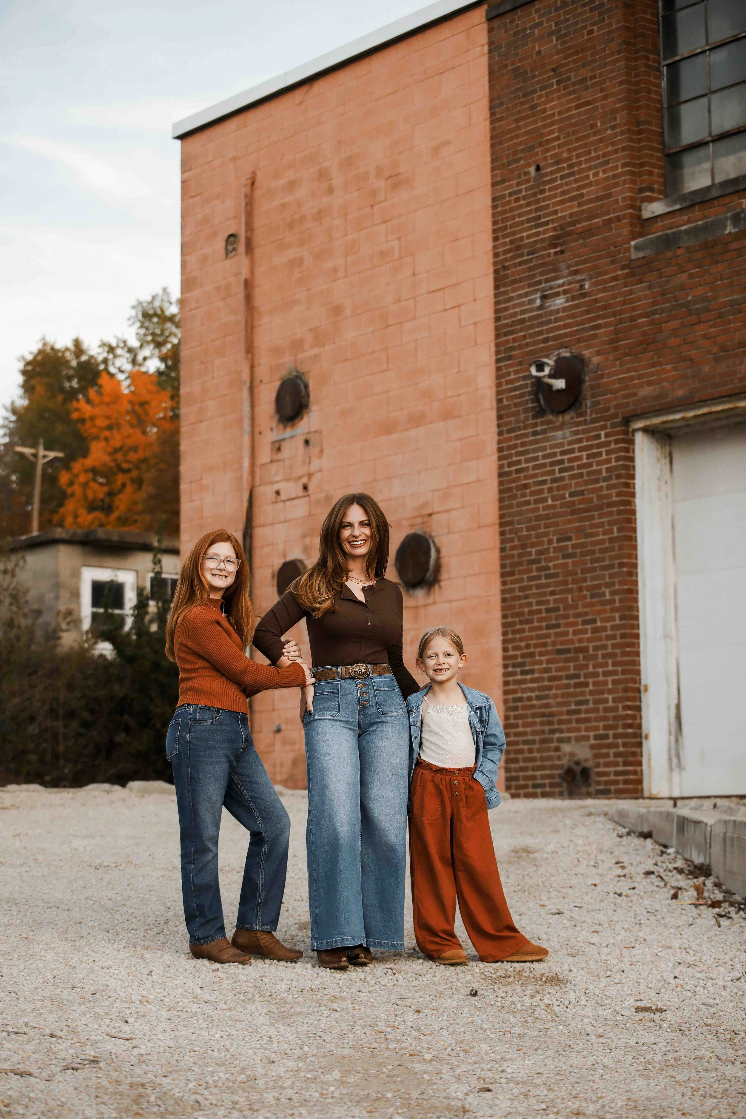 Casey Scott and her two young daughters standing outside in front of a brick building with autumn trees in the background, smiling at the camera.