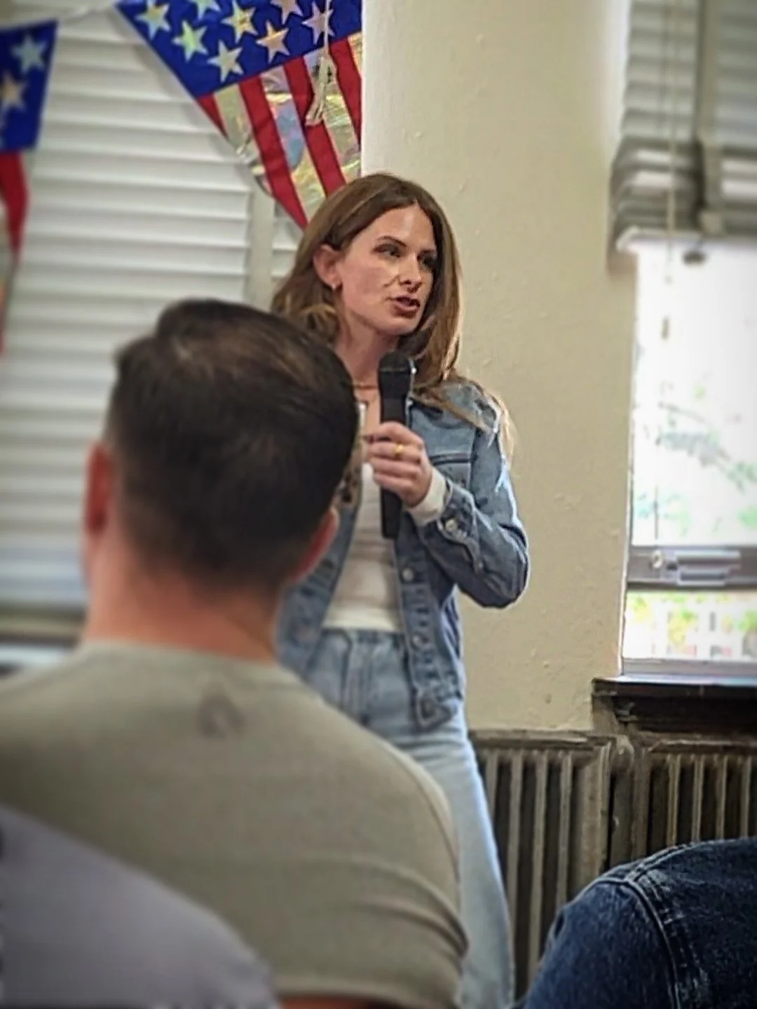 Casey Scott holding a microphone, speaking to a group of people, with red, white, and blue American flag decorations hanging in the background.