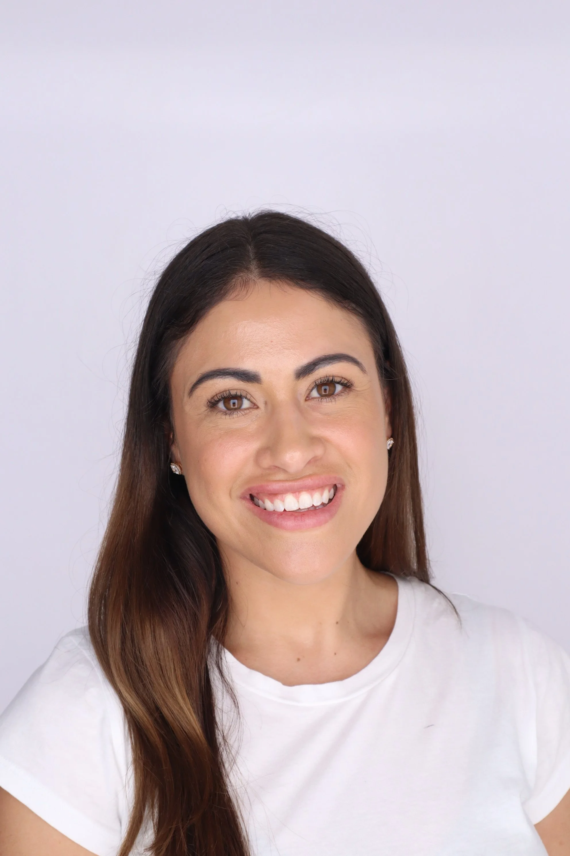 Close-up of a woman smiling, with long brown hair, light skin, wearing small earrings and a white t-shirt, standing against a plain light background.