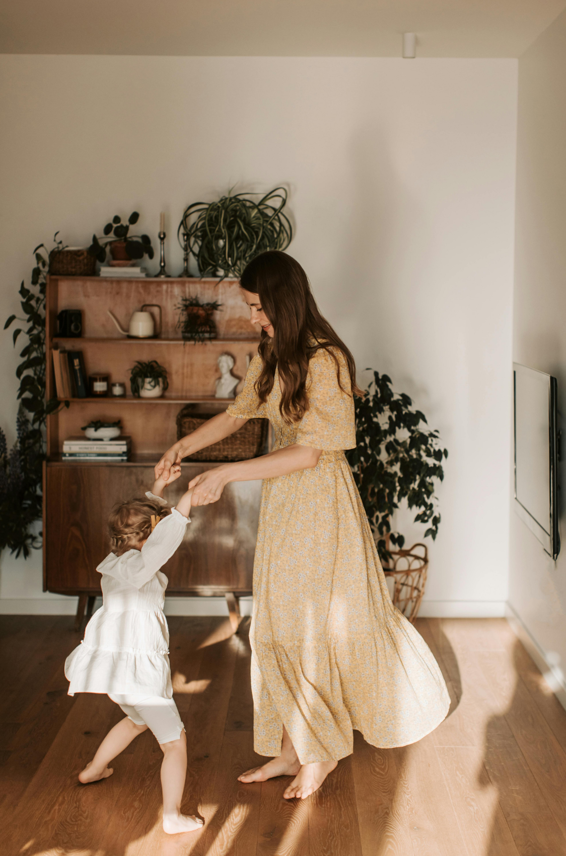 A woman and a young girl dancing together in a living room. The woman is wearing a yellow floral dress and the girl is wearing a white dress. The woman is smiling and holding the girl's hands, and they are spinning around on a wooden floor.
