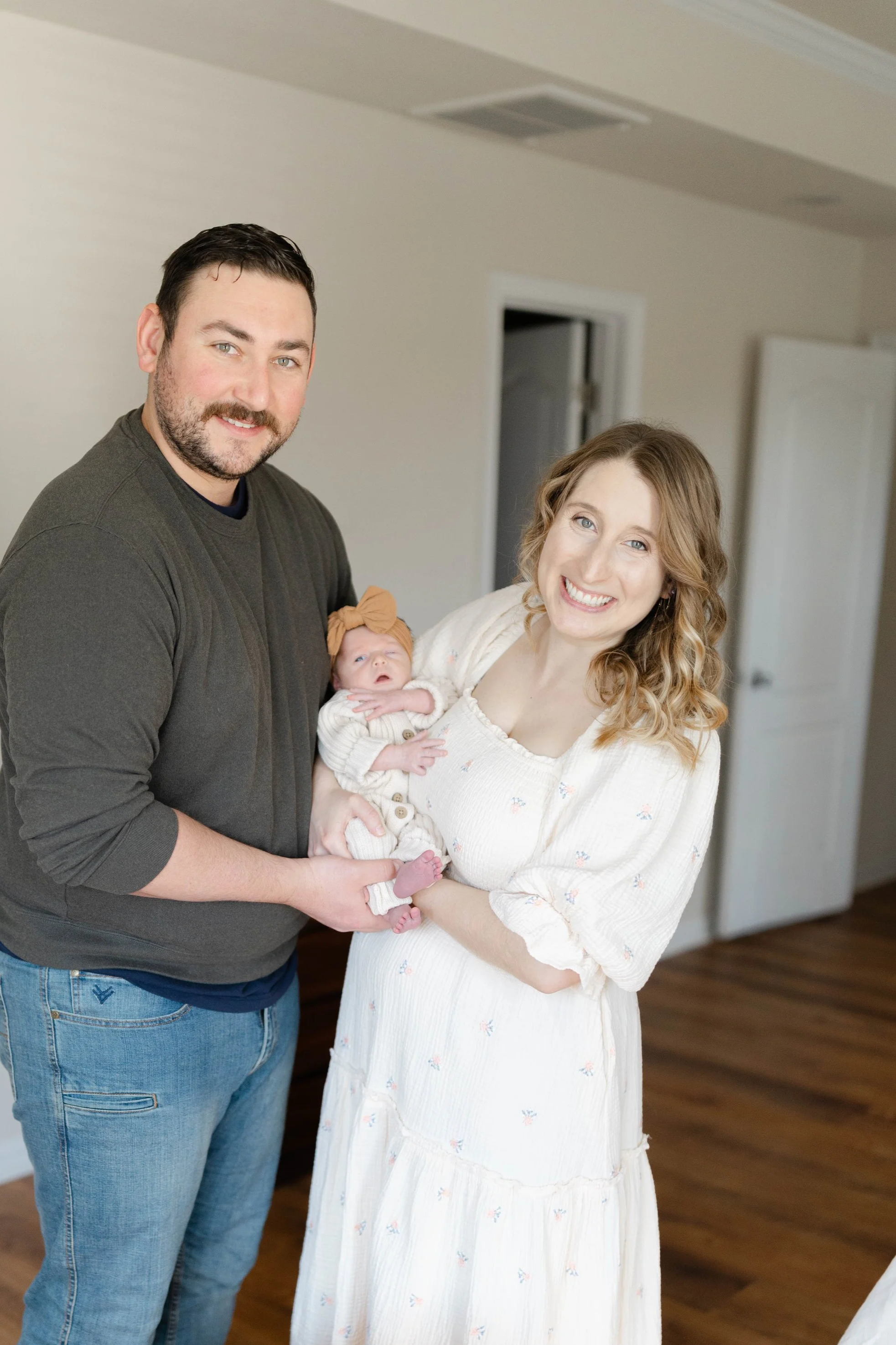 A smiling family of three standing together indoors; a man, a woman, and a baby girl held by the woman; all facing the camera.