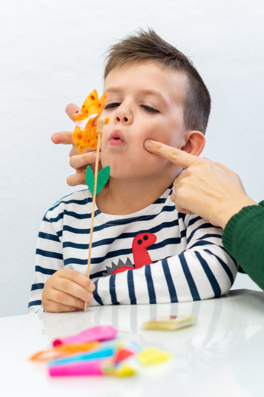 A young boy with short, styled hair wears a striped shirt with a red dinosaur patch. An adult's hand is gently pinching his cheek while he is looking at a paper butterfly on a stick in front of him. The scene appears to be indoors with a white background and colorful craft supplies on the table.