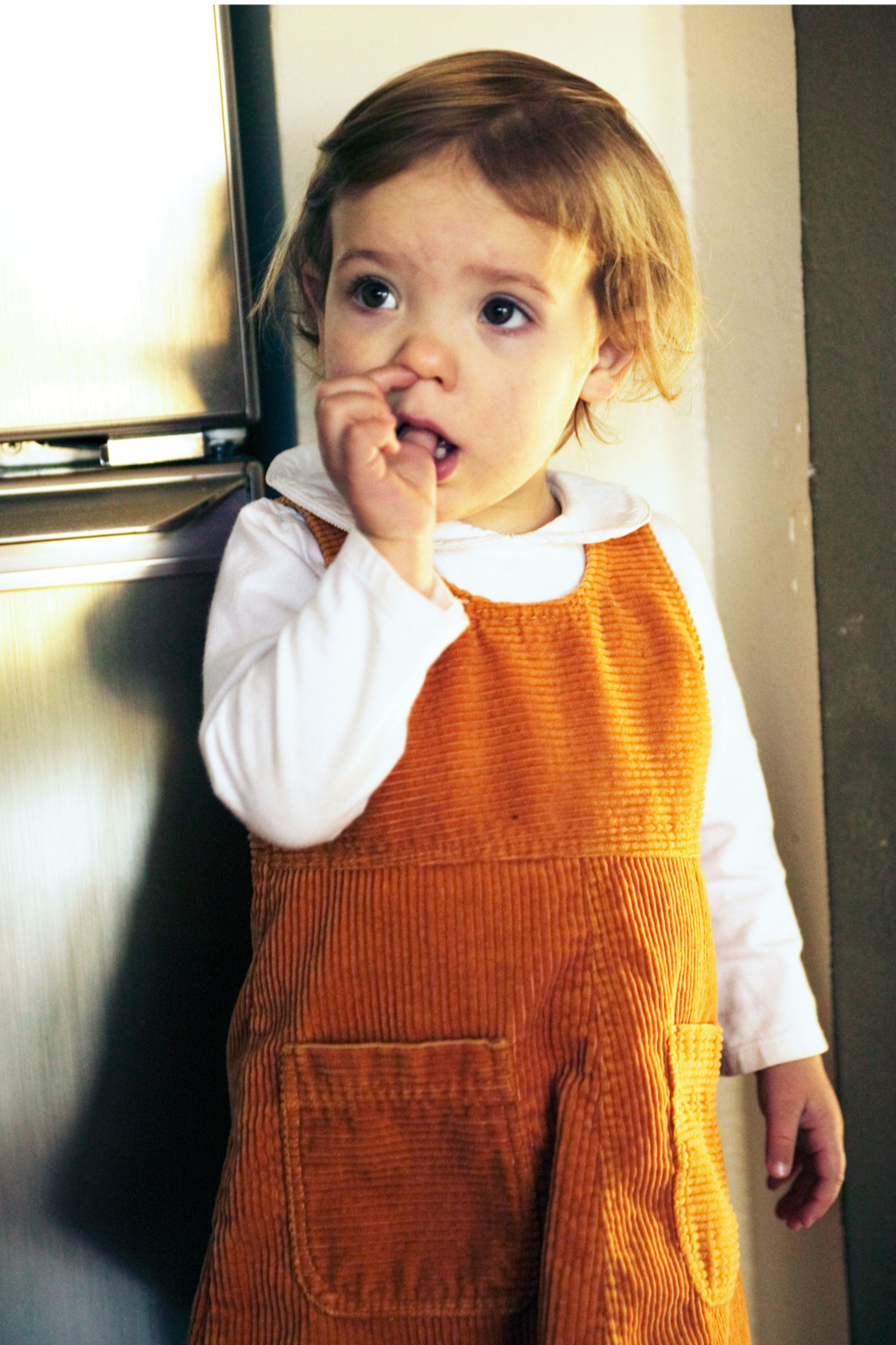 A young girl with short light brown hair and wide eyes, standing indoors by a metallic surface, wearing a white long-sleeve shirt and orange corduroy overalls, with her fingers near her mouth.