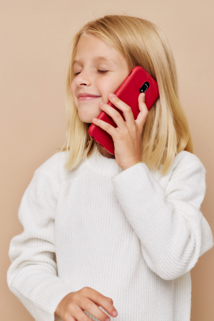 A young girl with blonde hair and a white sweater talking on a red mobile phone.