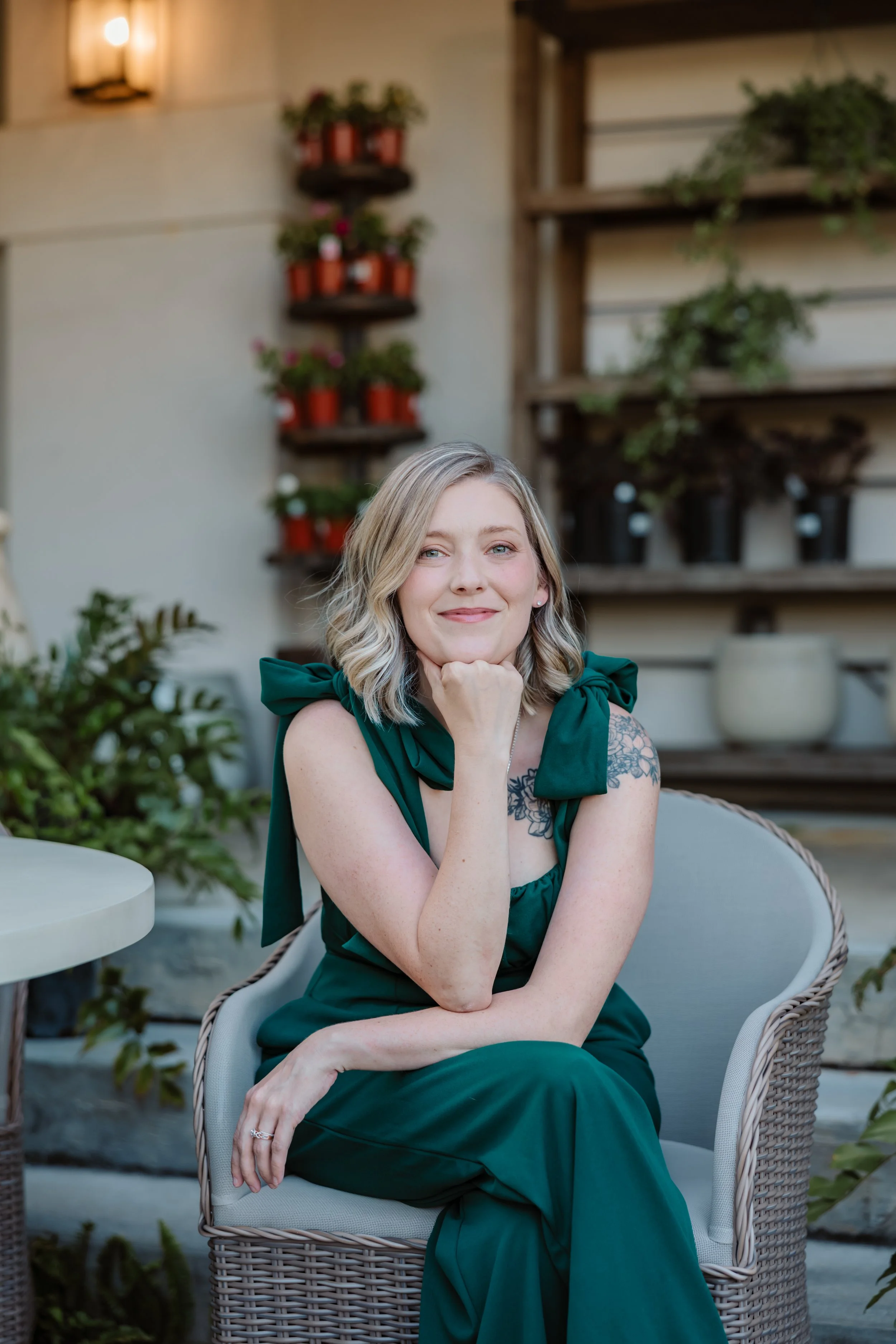 A woman with blonde hair, wearing a dark green dress with bows on the shoulders, sitting on a white wicker chair outdoors with greenery and potted plants in the background.