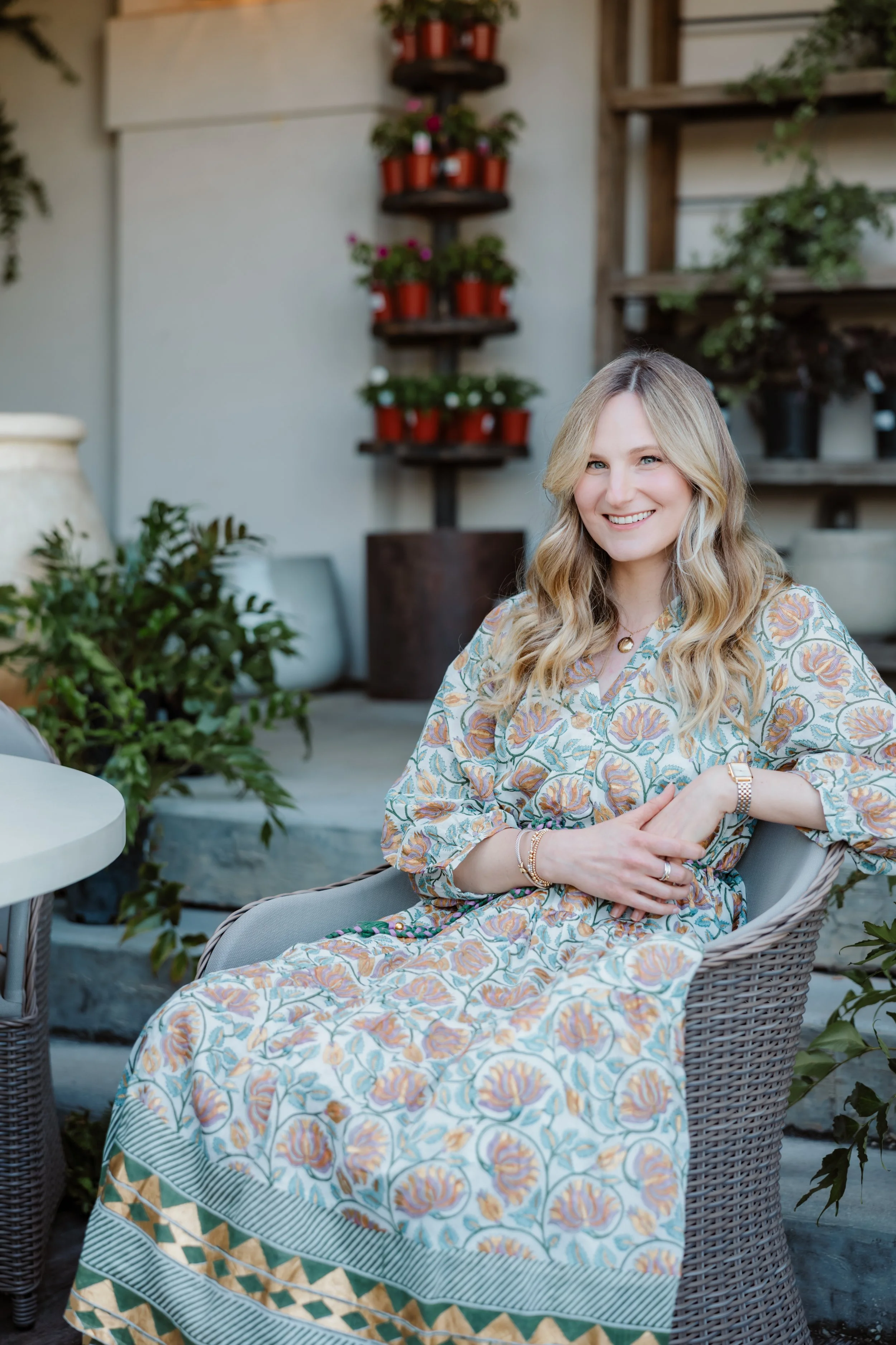 A woman with long blonde hair, smiling, wearing a floral dress, sitting in an outdoor furniture chair surrounded by plants and flowers in pots.