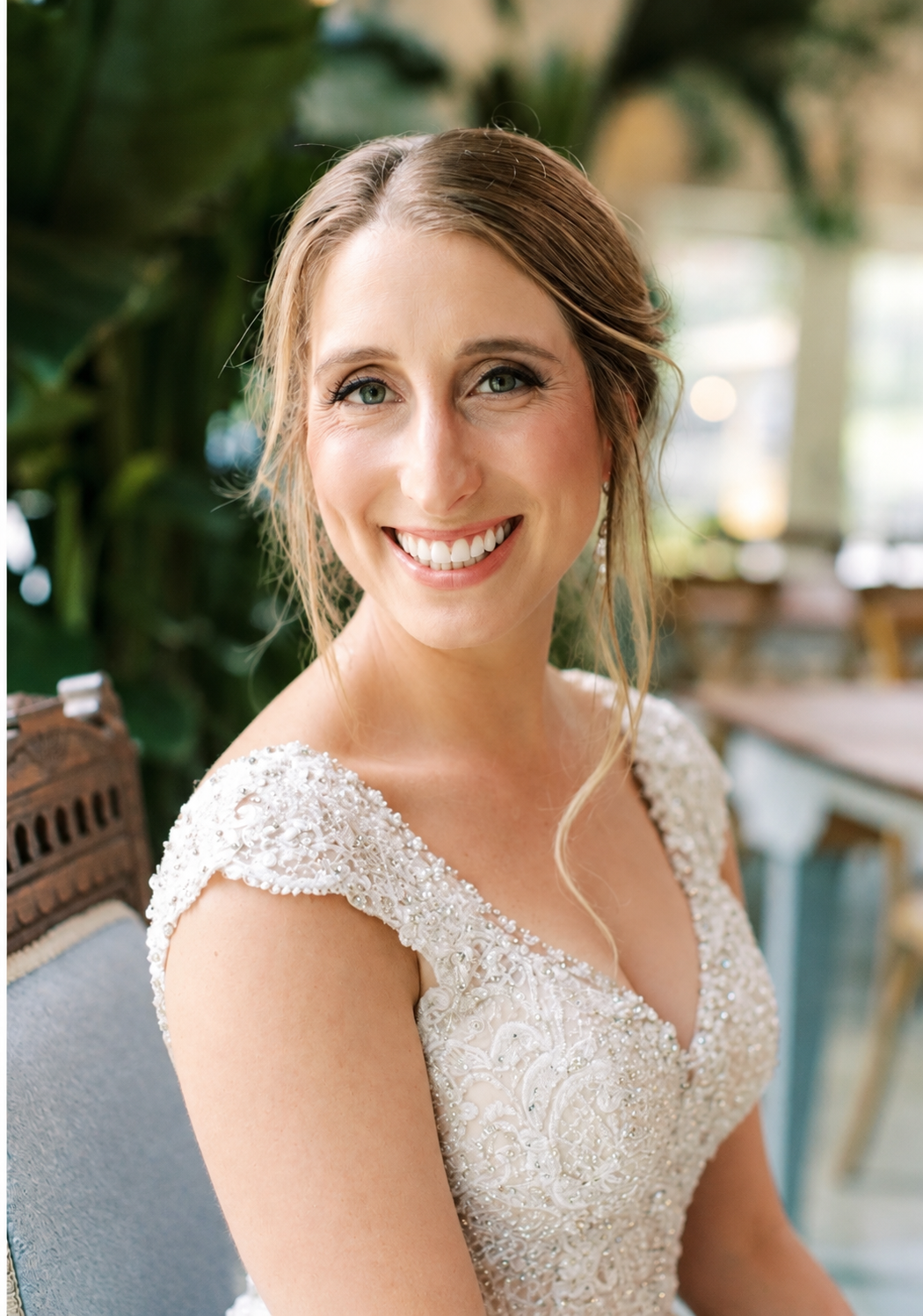 A smiling woman with light brown hair styled in loose waves, wearing a white beaded dress with short sleeves, sitting indoors with greenery and blurred furniture in the background.