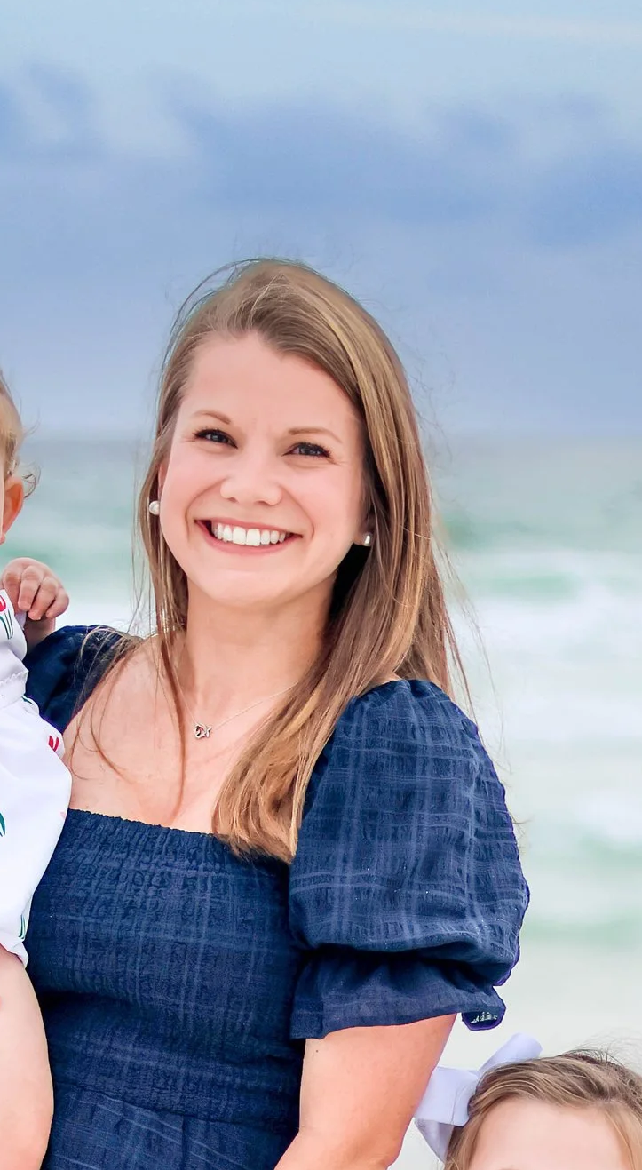 A smiling woman with long light brown hair at the beach, wearing a navy blue dress with puffed sleeves and pearl earrings, standing in front of ocean waves under a cloudy sky.