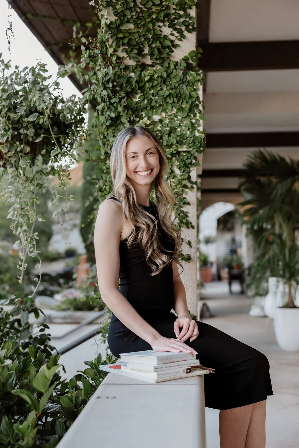 A young woman with long wavy blonde hair, wearing a black sleeveless dress, sitting on a white bench outdoors next to a wall covered with green ivy. She is smiling at the camera and has a stack of books in front of her.
