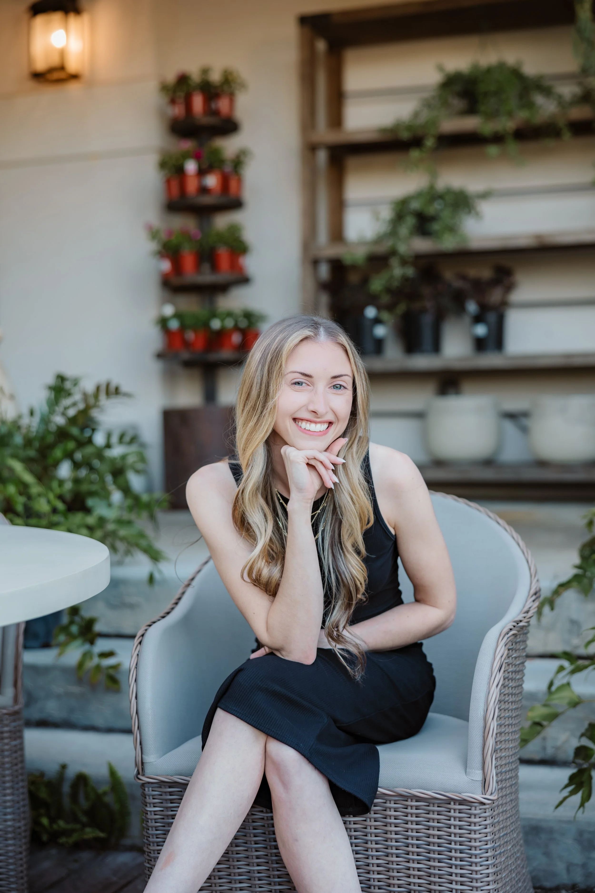 A young woman with long blond hair, smiling and resting her chin on her hand, sitting on a wicker chair in an outdoor setting with plants and a wall-mounted shelf with potted plants in the background.