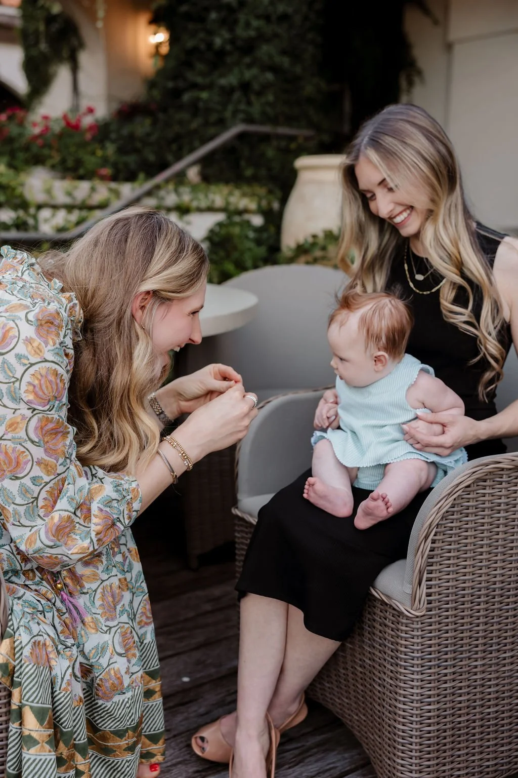 Two women and a baby girl sitting outside, with one woman leaning forward and the other holding the baby, all smiling and engaging with each other.