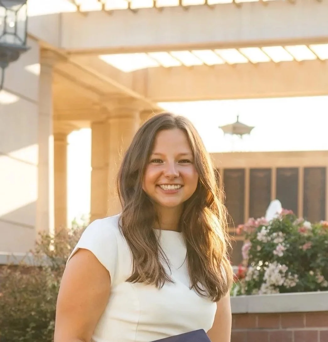A woman smiling outdoors with sunlight behind her, standing in front of flowering bushes and a wooden structure.