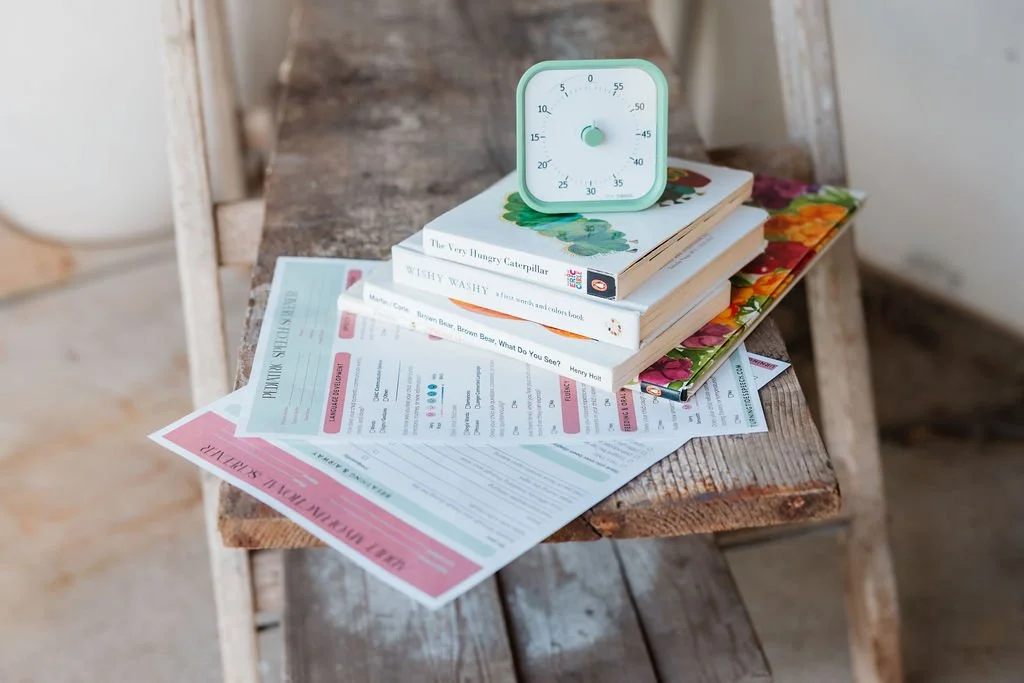 A wooden chair with a stack of books, papers, and a tea timer. The papers include a worksheet with pink headers.
