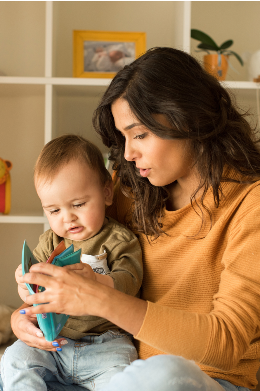 A woman and a young child sitting together, looking at a colorful book. The woman appears to be reading or explaining the book to the child, in a cozy indoor setting.