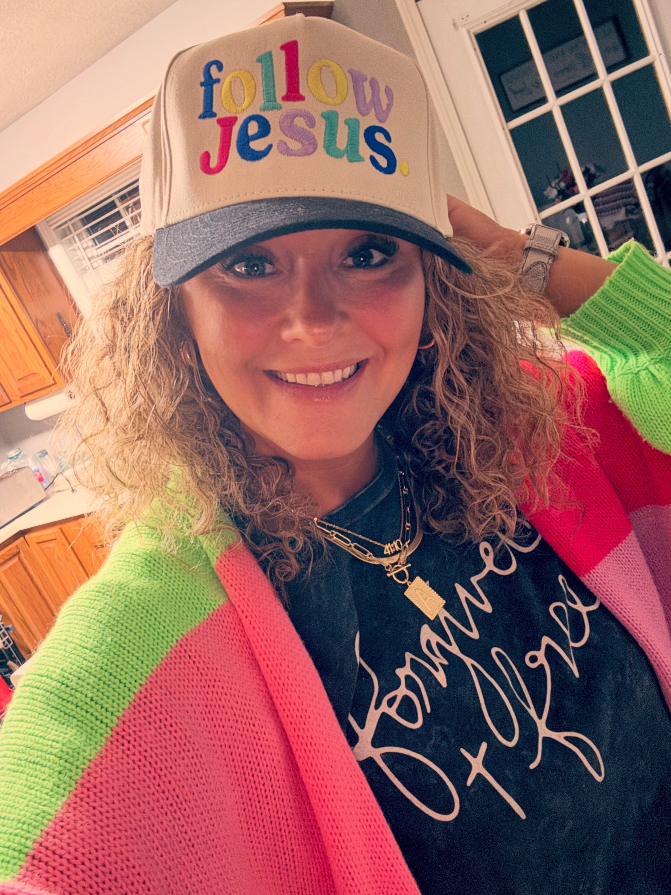 A woman with curly hair smiling at the camera, wearing a baseball cap with colorful embroidered text that says 'follow Jesus,' a black T-shirt, and colorful neon green and pink sweater.