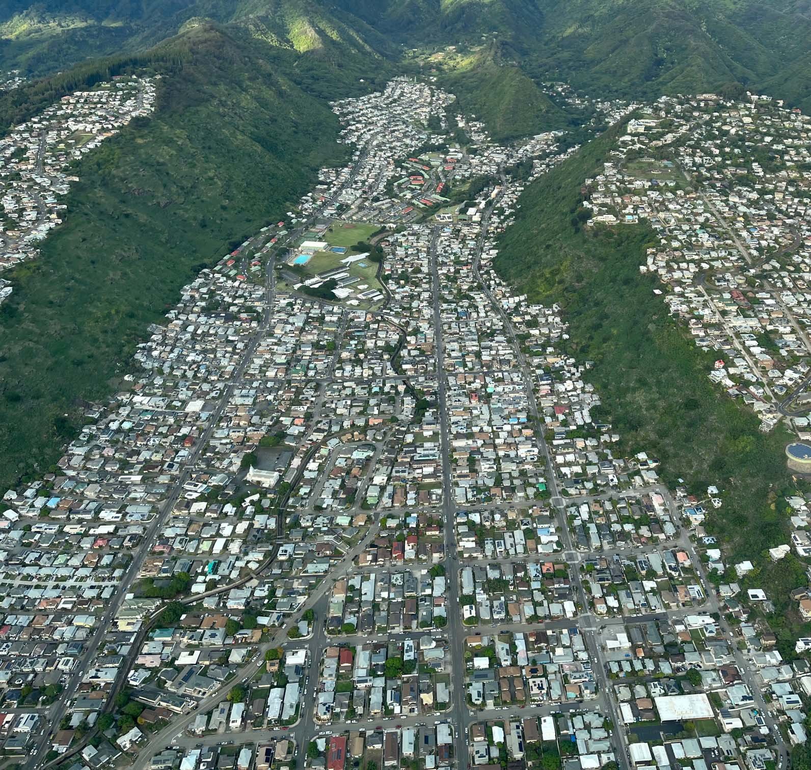 An aerial view of a residential neighborhood situated in a valley surrounded by green mountains, featuring a dense layout of houses, roads, and some recreational facilities.