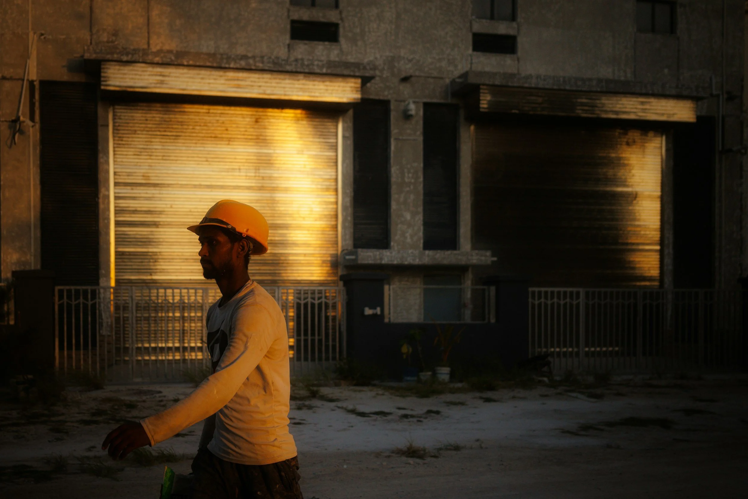 A man wearing an orange hard hat and a white long-sleeve shirt sitting on a tree stump near a residential building with metal shutters at sunset.
