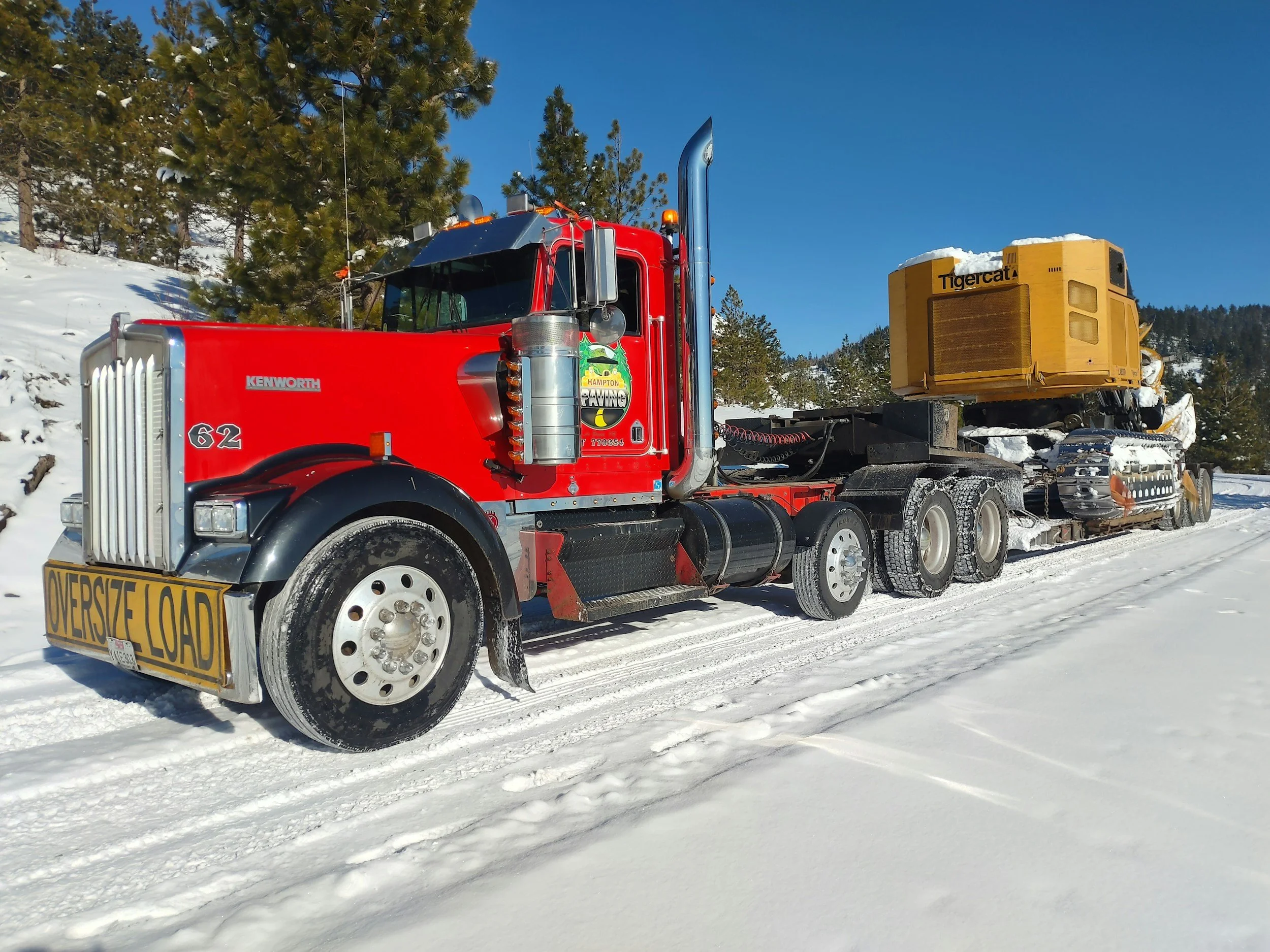 Red semi-truck with a yellow oversize load sign on the front, carrying a large yellow construction or industrial machine, driving on snowy mountainous terrain with trees and blue sky in the background.