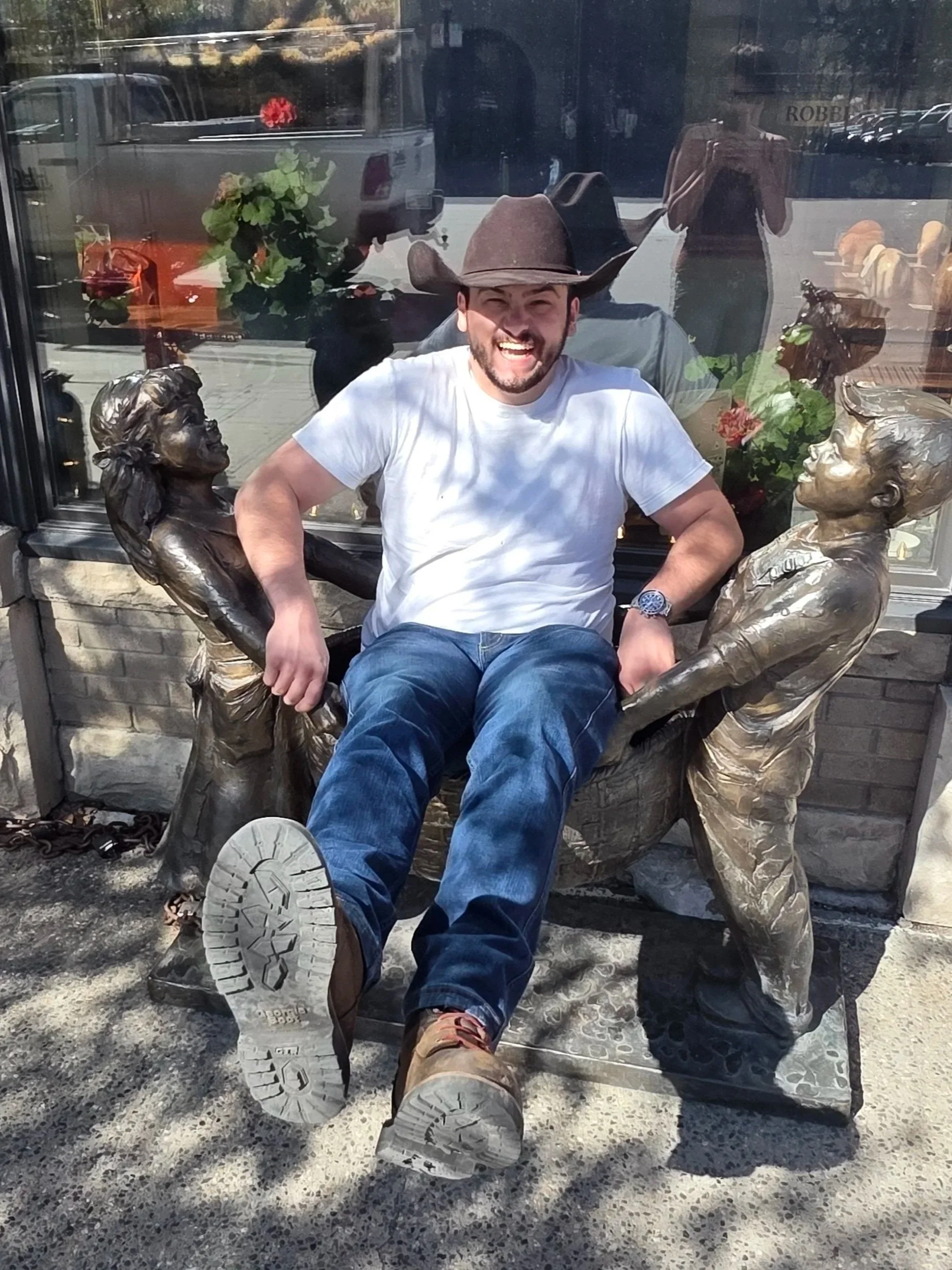 A man wearing a cowboy hat, white t-shirt, and jeans sitting on a bronze sculpture bench with two women sculptures on either side, outside a storefront. The man is smiling with his mouth open, leg crossed, leaning back slightly, and wearing a wristwa