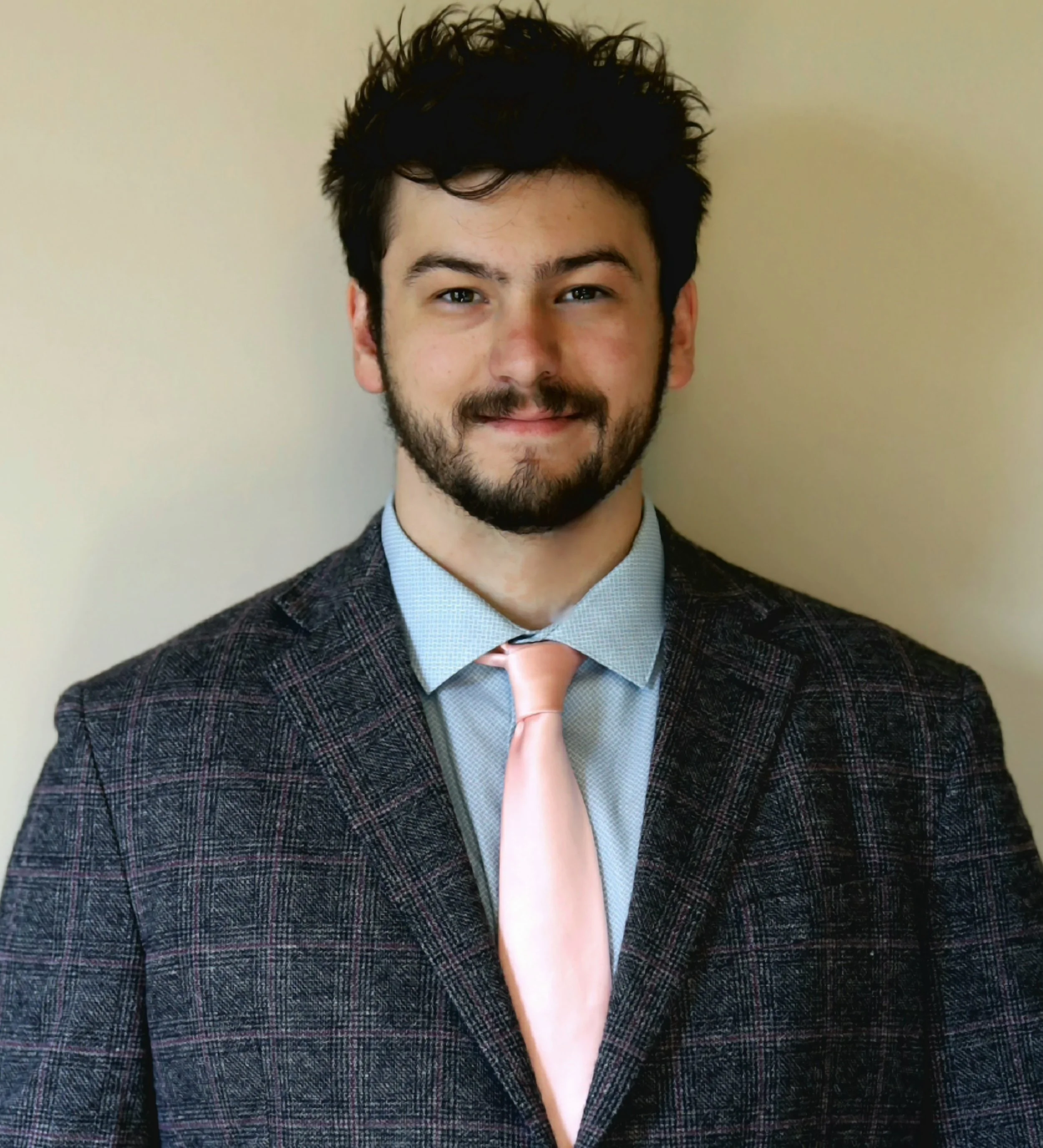 A young man with dark messy hair, smiling, dressed in a plaid blazer, light blue shirt, and pink tie, standing against a beige wall.