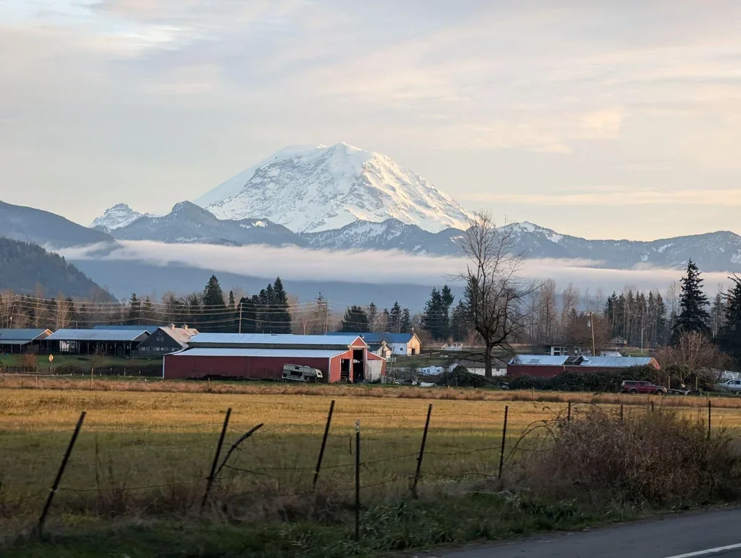 A rural farm scene with a large snow-capped mountain in the background, likely Mount Rainier. The foreground shows fields, a red barn, and farm buildings with a partly cloudy sky.