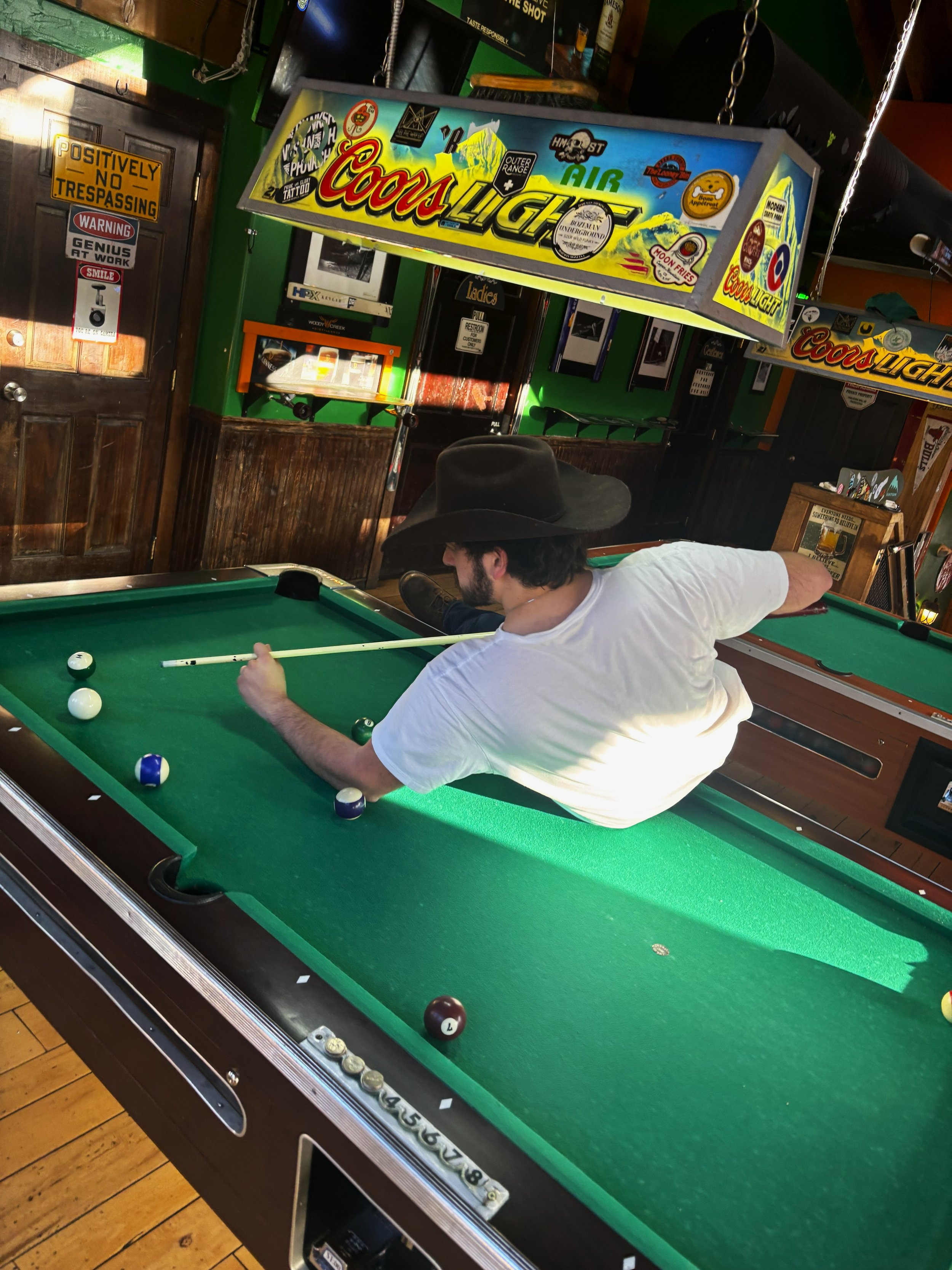 A man wearing a wide-brimmed black hat and a white T-shirt playing pool at a bar or game room.