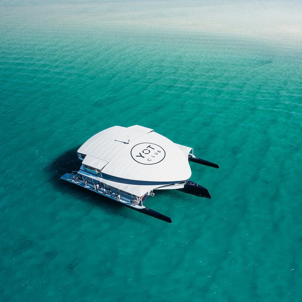 A catamaran boat with a white canopy labeled "YOT CLUB" floats on turquoise water.