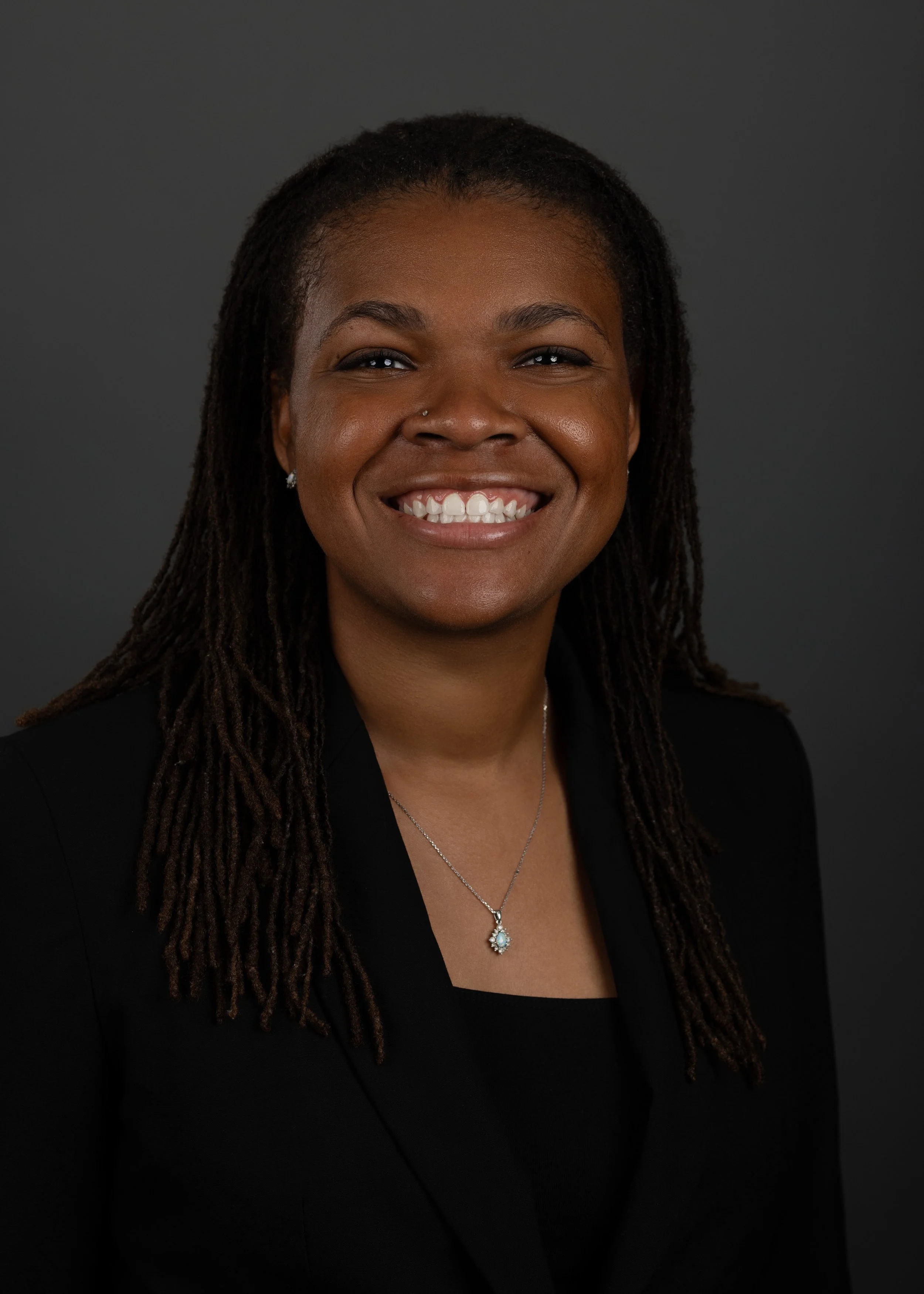 Headshot of a smiling woman with dark skin, long dreadlocks, a nose piercing, wearing earrings, a necklace with a pendant, and a black blazer against a dark background.