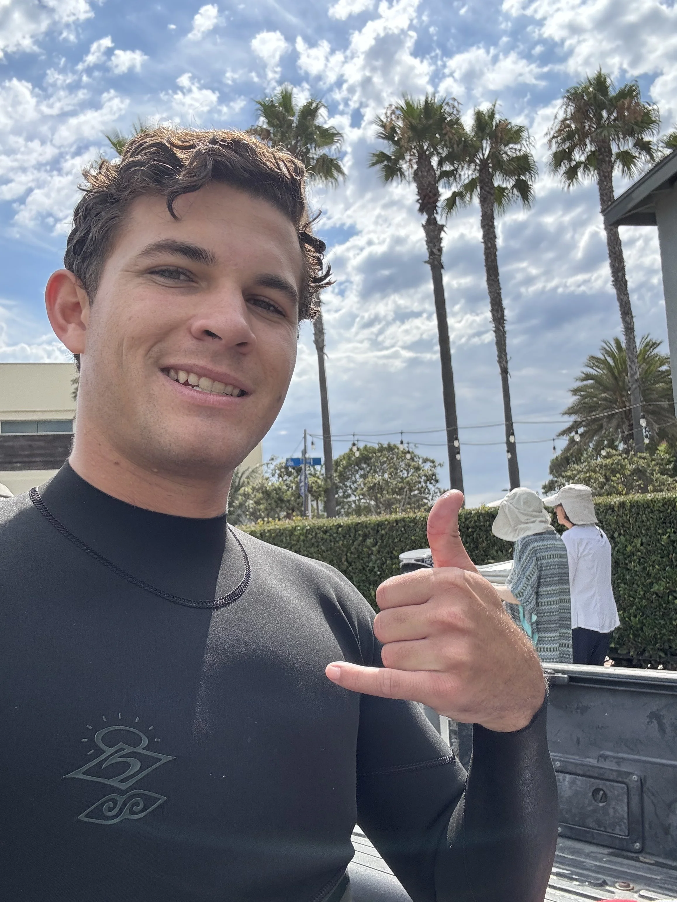 Young man smiling, wearing a black athletic shirt with a logo, giving a thumbs-up, with palm trees and a partly cloudy sky in the background.