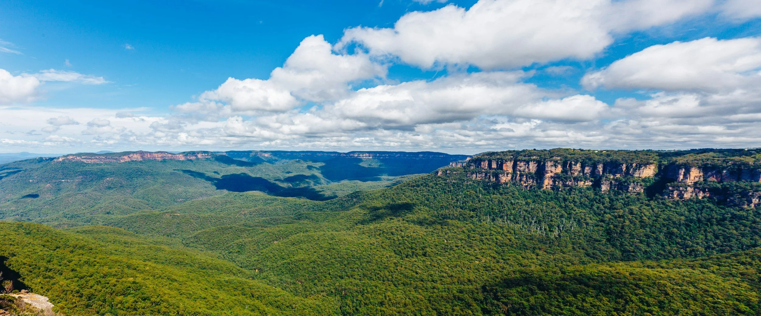Scenic view of forested mountains with rocky cliffs on the right, under a partly cloudy blue sky.