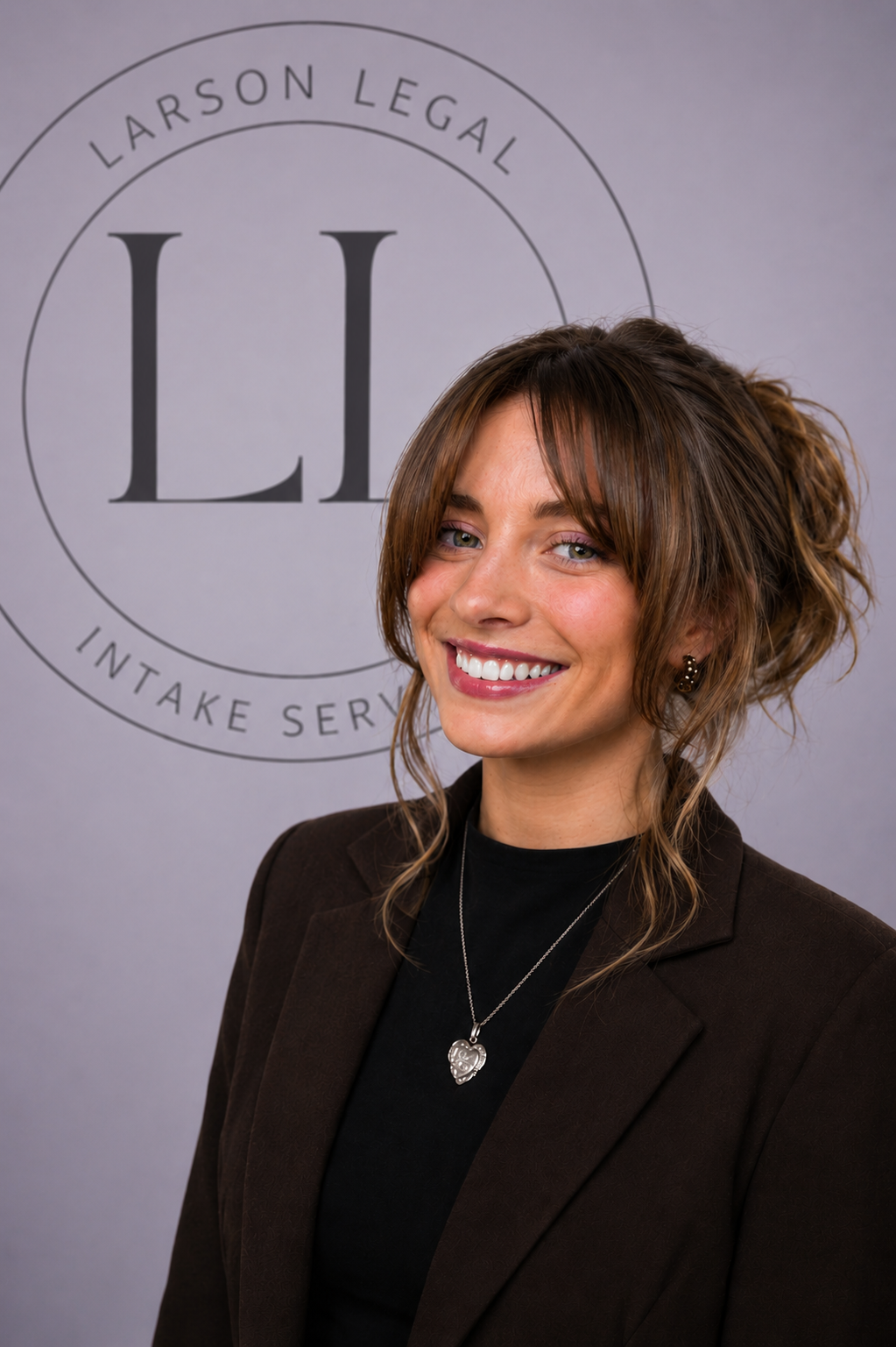 A woman smiling with brown hair styled in loose curls, wearing a black top, a dark blazer, and a necklace with a heart-shaped pendant, standing in front of a gray backdrop with the logo 'Larson Legal Intake Services'.