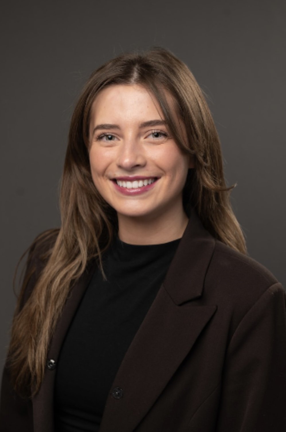 Portrait of a smiling woman with long brown hair, wearing a black top and dark blazer against a gray background.