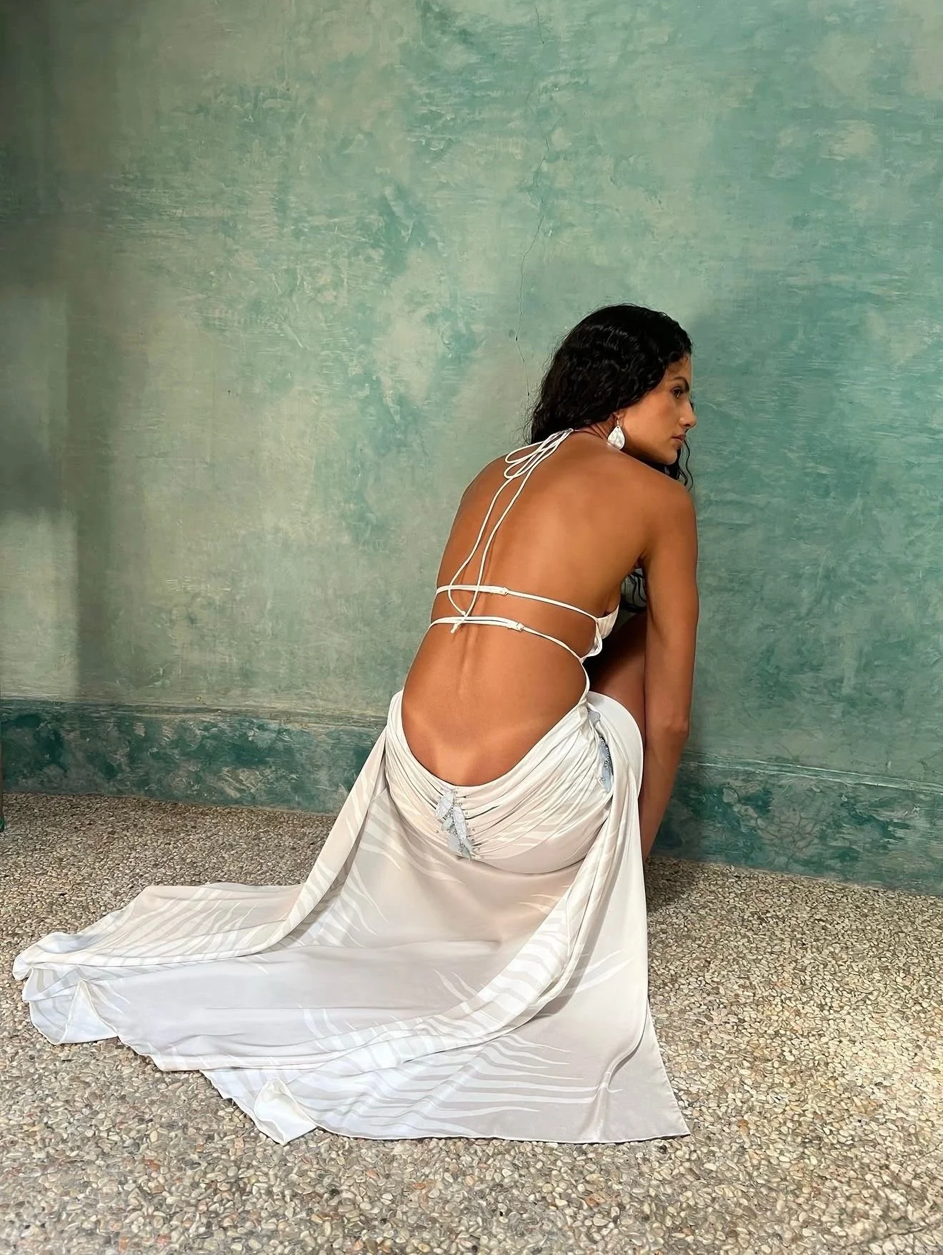 Model in a white backless gown seated against a green plaster wall