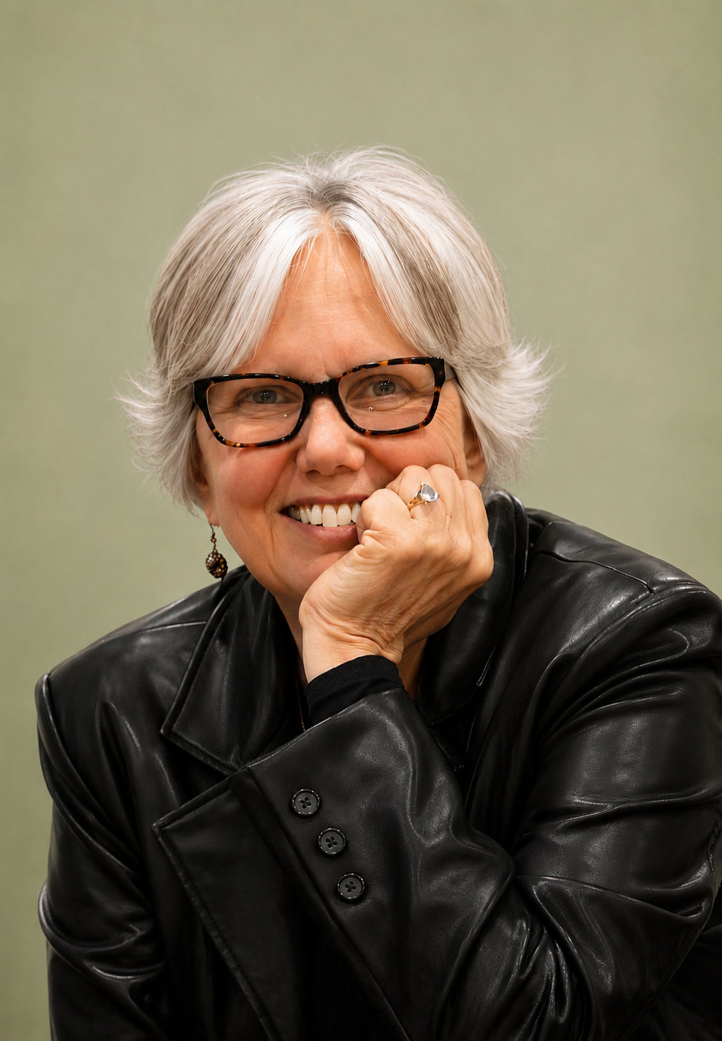 Smiling woman with short gray hair, glasses, and earrings, wearing a black leather jacket, resting her chin on her hand, against a plain green background.